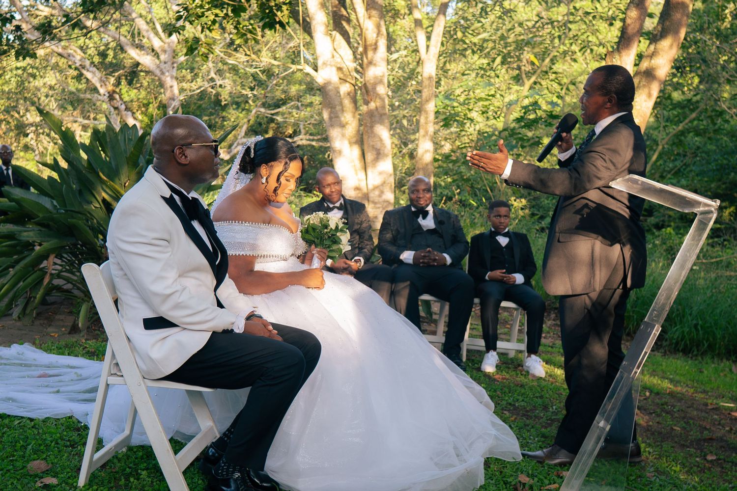 Outdoor wedding ceremony with bride and groom seated before officiant, groomsmen in background, lush green trees.