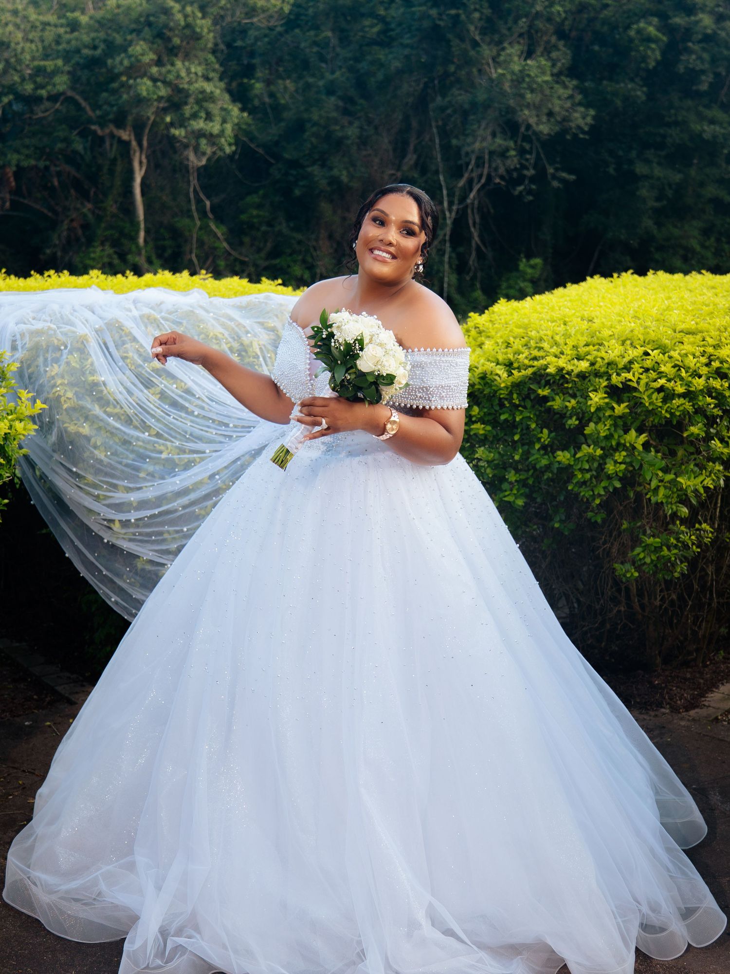 Bride in white off-shoulder ball gown with flowing veil holds bouquet outdoors near lush green hedges.