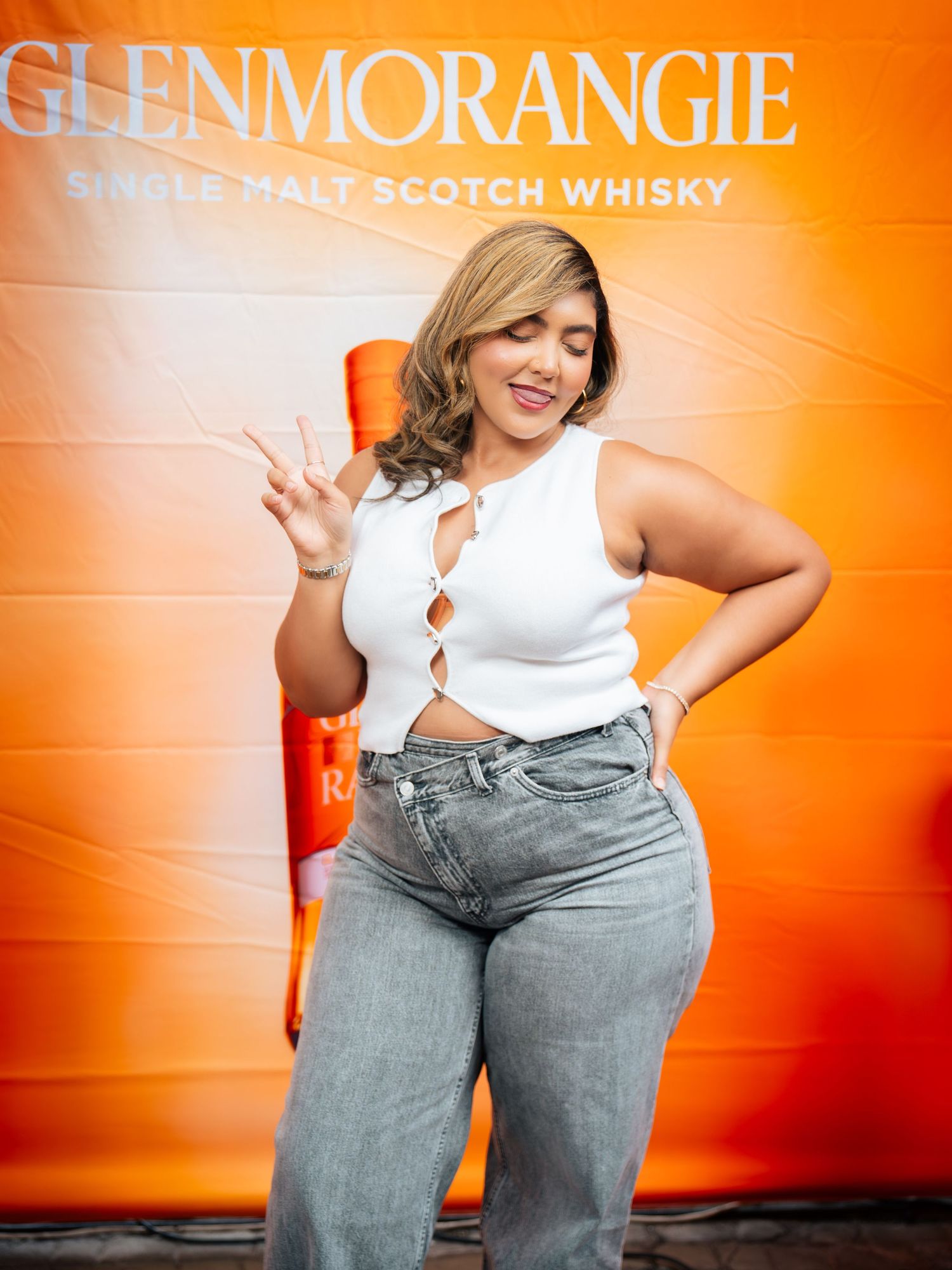 Woman in white crop top and jeans poses with peace sign at Glenmorangie Single Malt Scotch Whisky event.