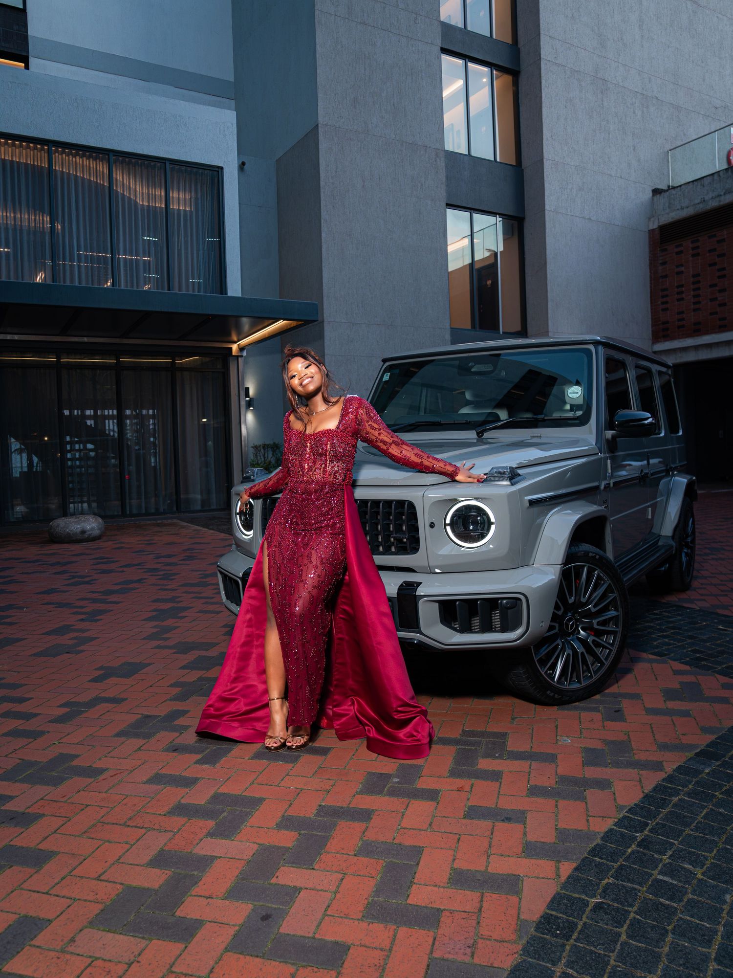 Woman in red gown poses beside silver Mercedes-Benz G-Class SUV on brick driveway outside modern building.