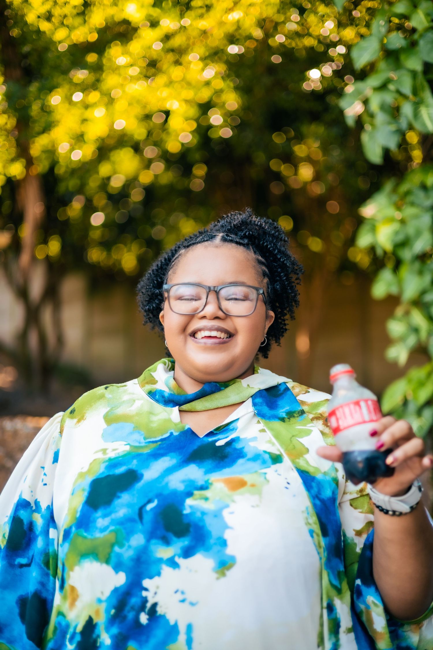 Smiling woman in colorful tie-dye top holding a Coca-Cola bottle outdoors with green trees in background.