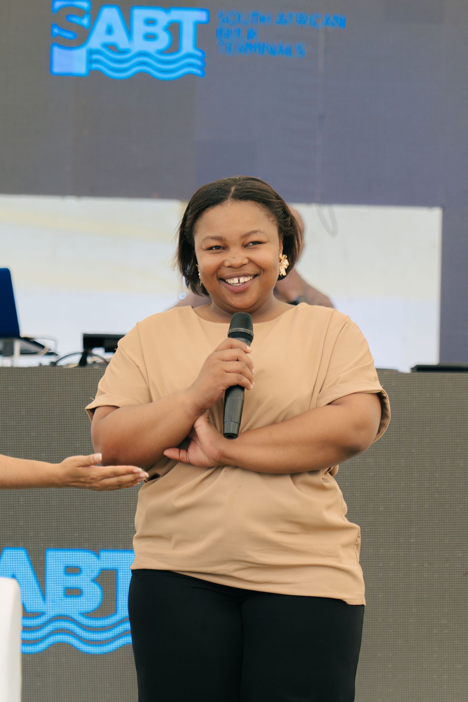 Smiling woman holding microphone on stage at SABT South African event wearing beige t-shirt.
