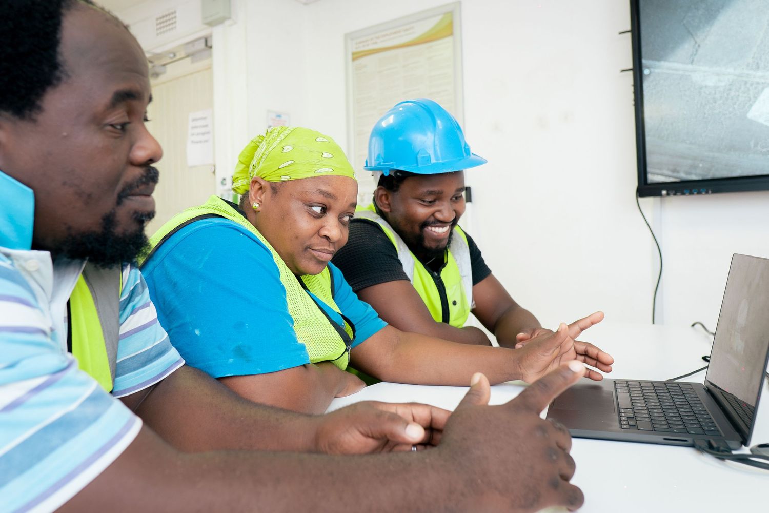 Three construction workers in safety vests and hard hats collaborate over a laptop in an office setting.