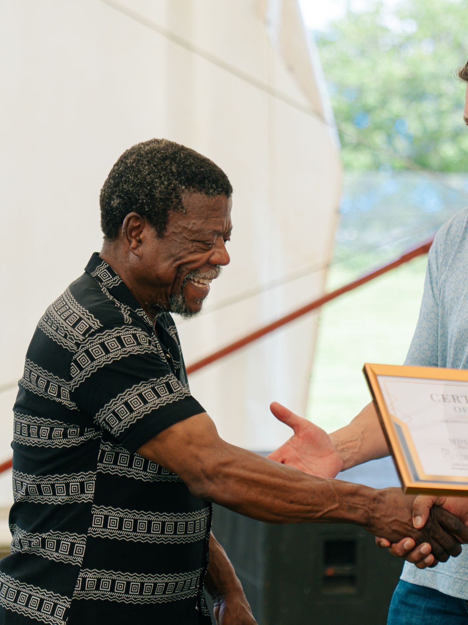 Smiling man in striped polo shirt shaking hands and receiving a framed certificate outdoors.