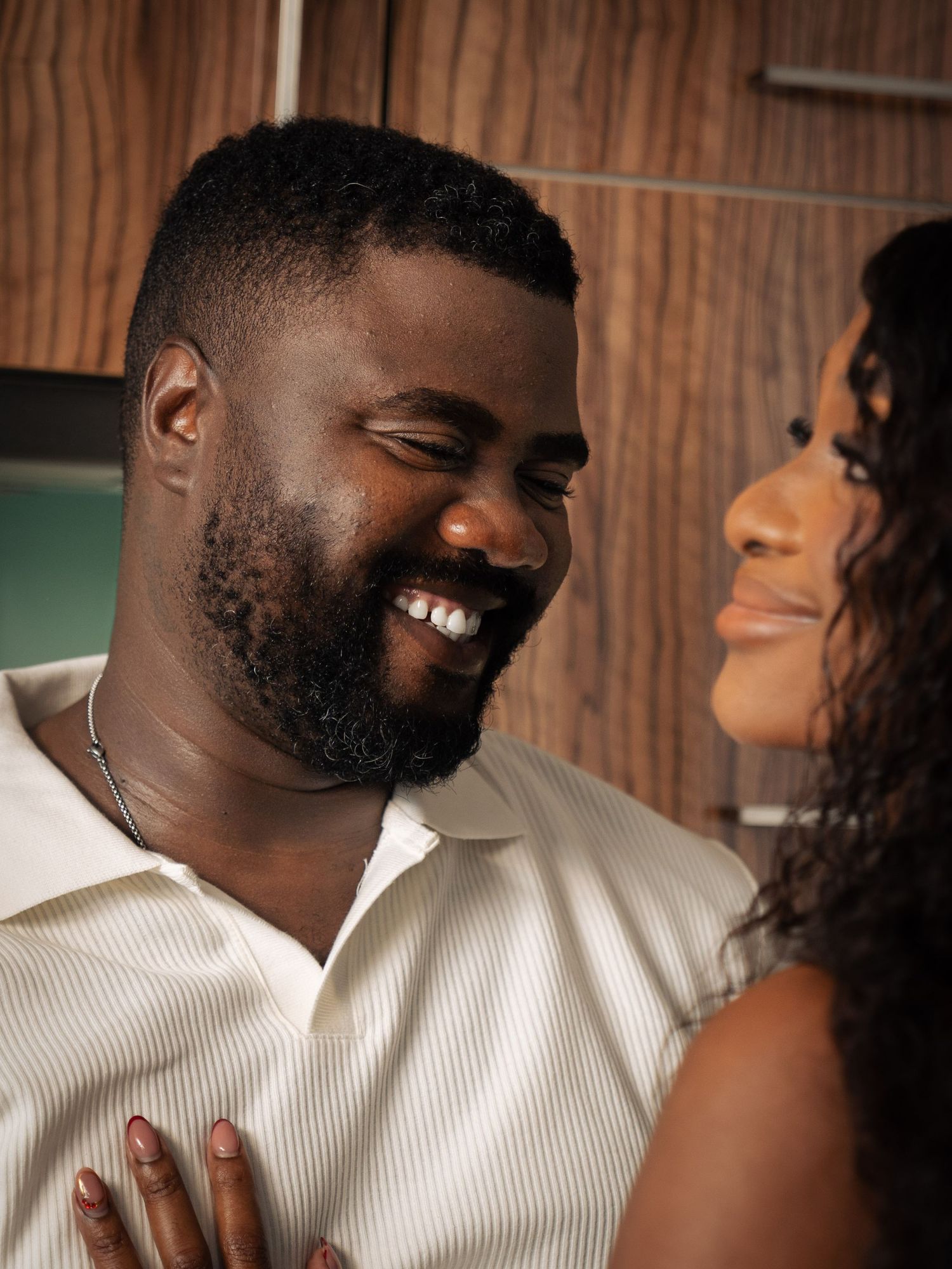 A smiling Black man with a beard laughs joyfully while facing a woman in a warmly lit indoor setting.