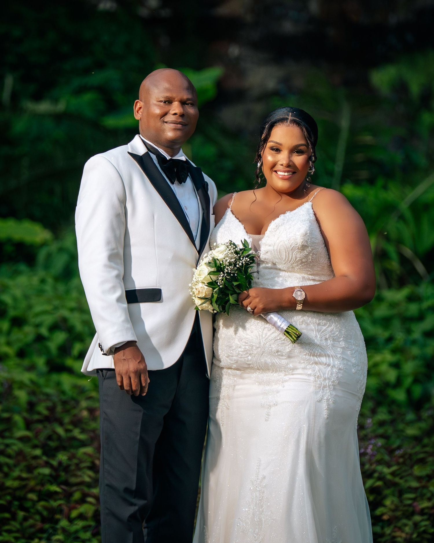 Couple posing in wedding attire, groom in white tuxedo and bride in white gown holding bouquet, outdoors.