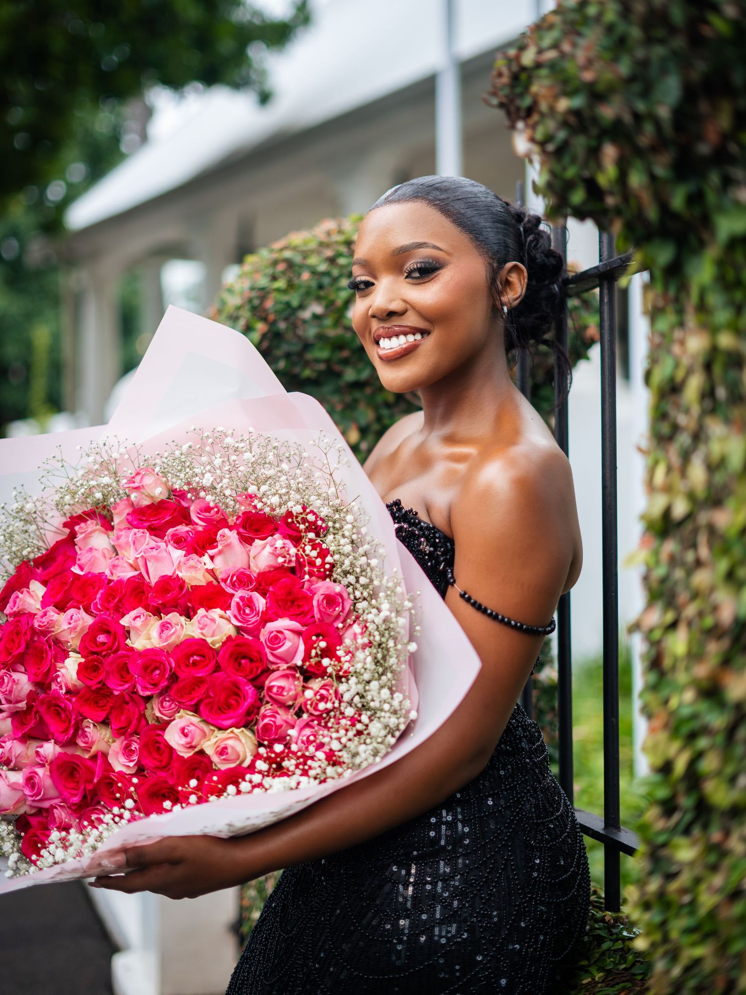 Smiling young Black woman in black sequin dress holding large bouquet of pink and red roses.