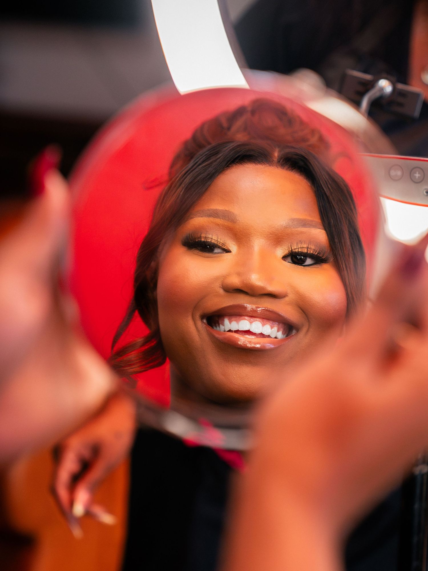 A smiling woman with red hair and glam makeup is reflected in a round mirror during a professional makeup session.