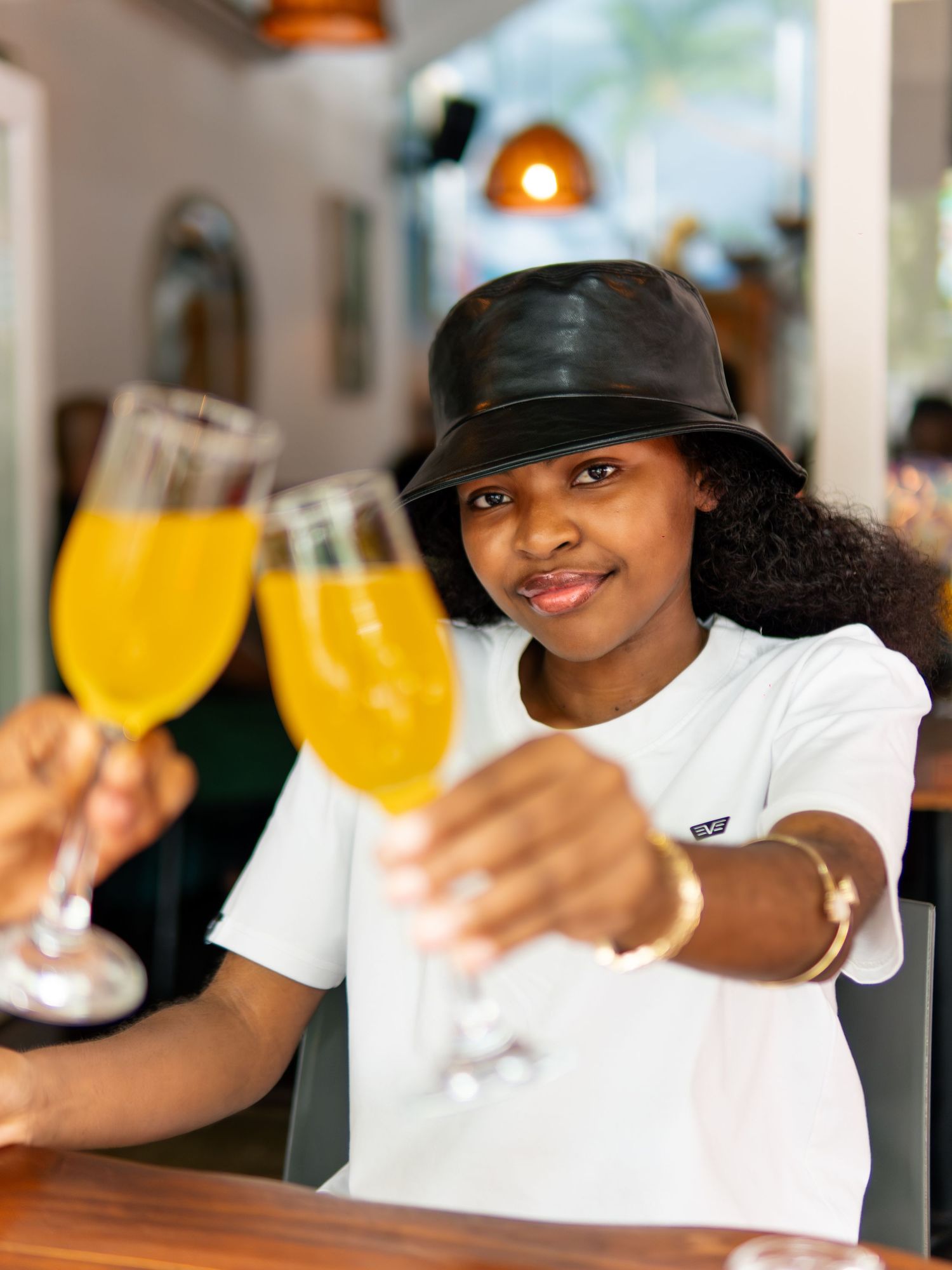 Woman in black bucket hat toasting with mimosas at a bright restaurant bar, smiling and holding champagne flutes.
