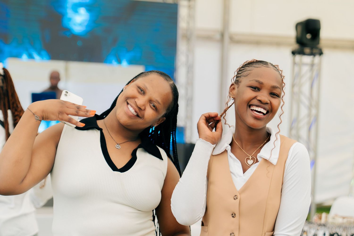 Two young women smiling and posing together at an outdoor event, one in a white top and the other in a beige vest.