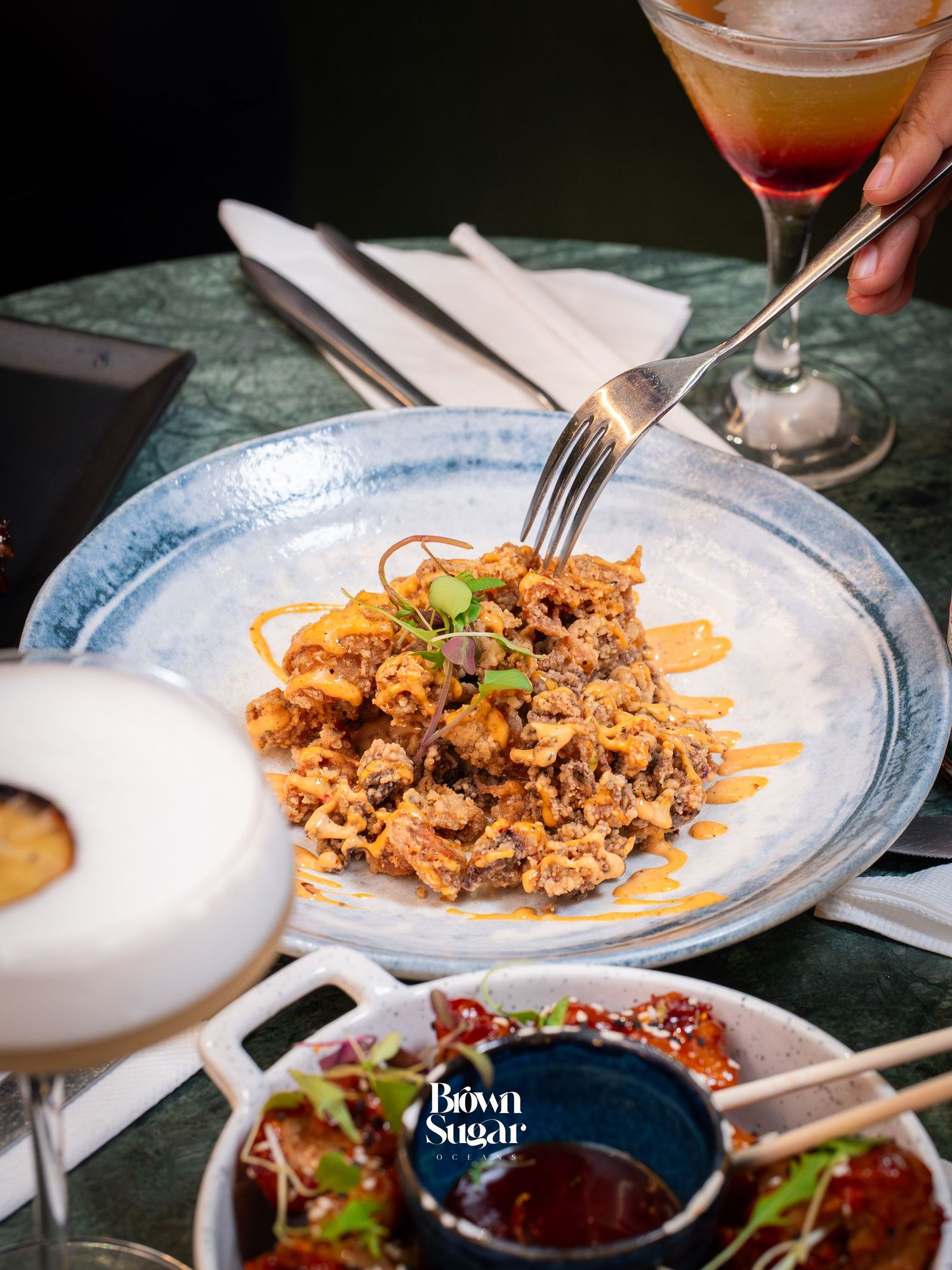 A fork rests in a spicy noodle dish on a blue plate at Brown Sugar restaurant, with cocktails and appetizers nearby.