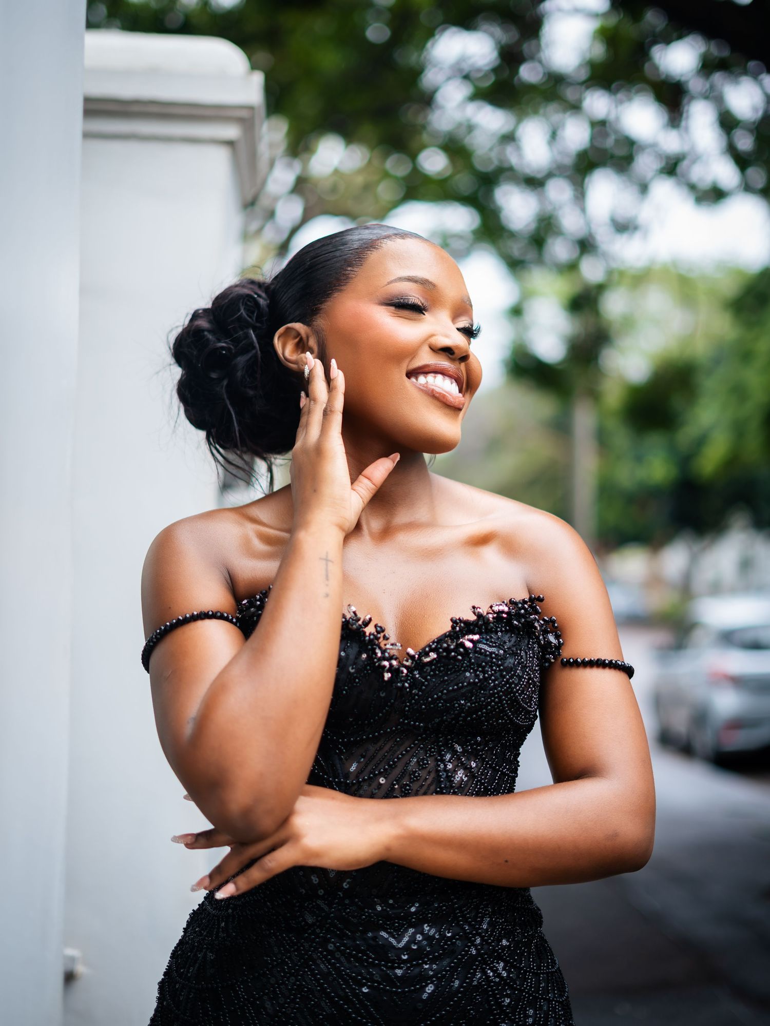A young Black woman in a black off-shoulder beaded gown smiles elegantly on a tree-lined street.