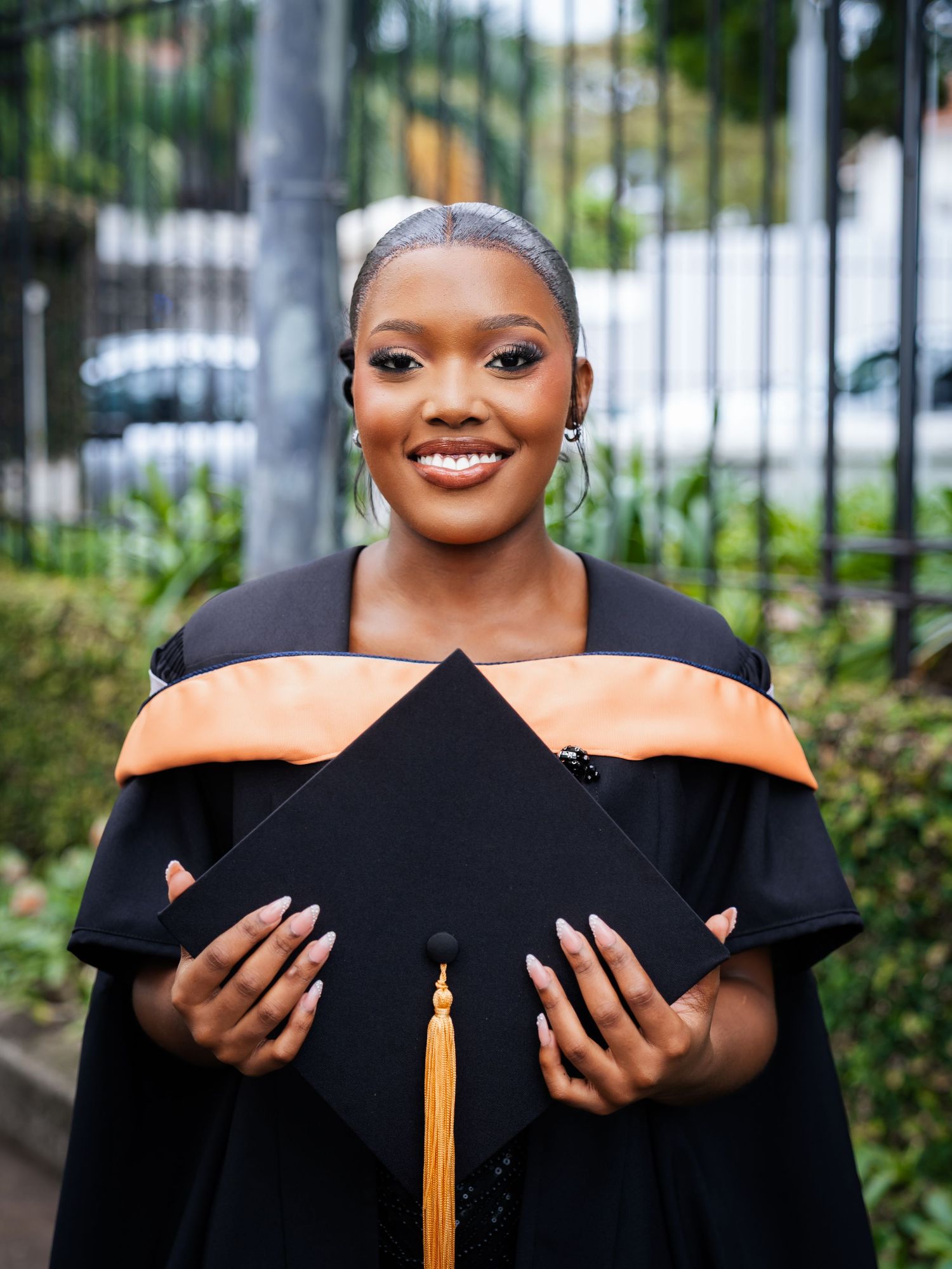 Smiling graduate in black and orange academic regalia holding mortarboard cap outdoors at graduation ceremony.