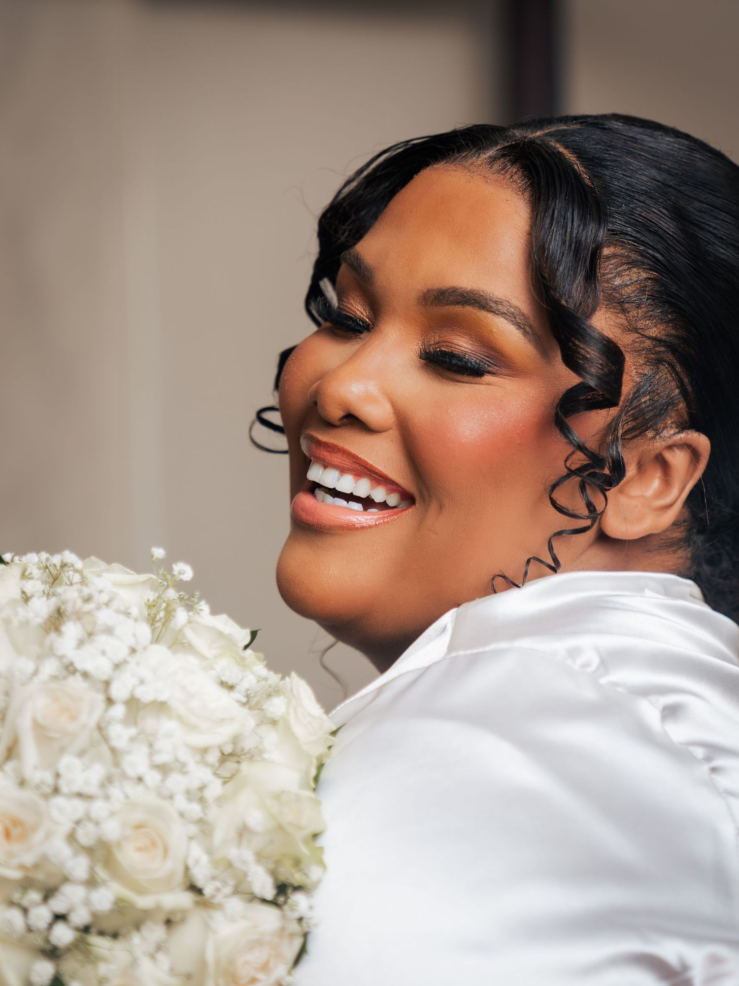 A radiant bride smiles holding a white rose bouquet, wearing a satin gown with elegant curled hair and glam makeup.