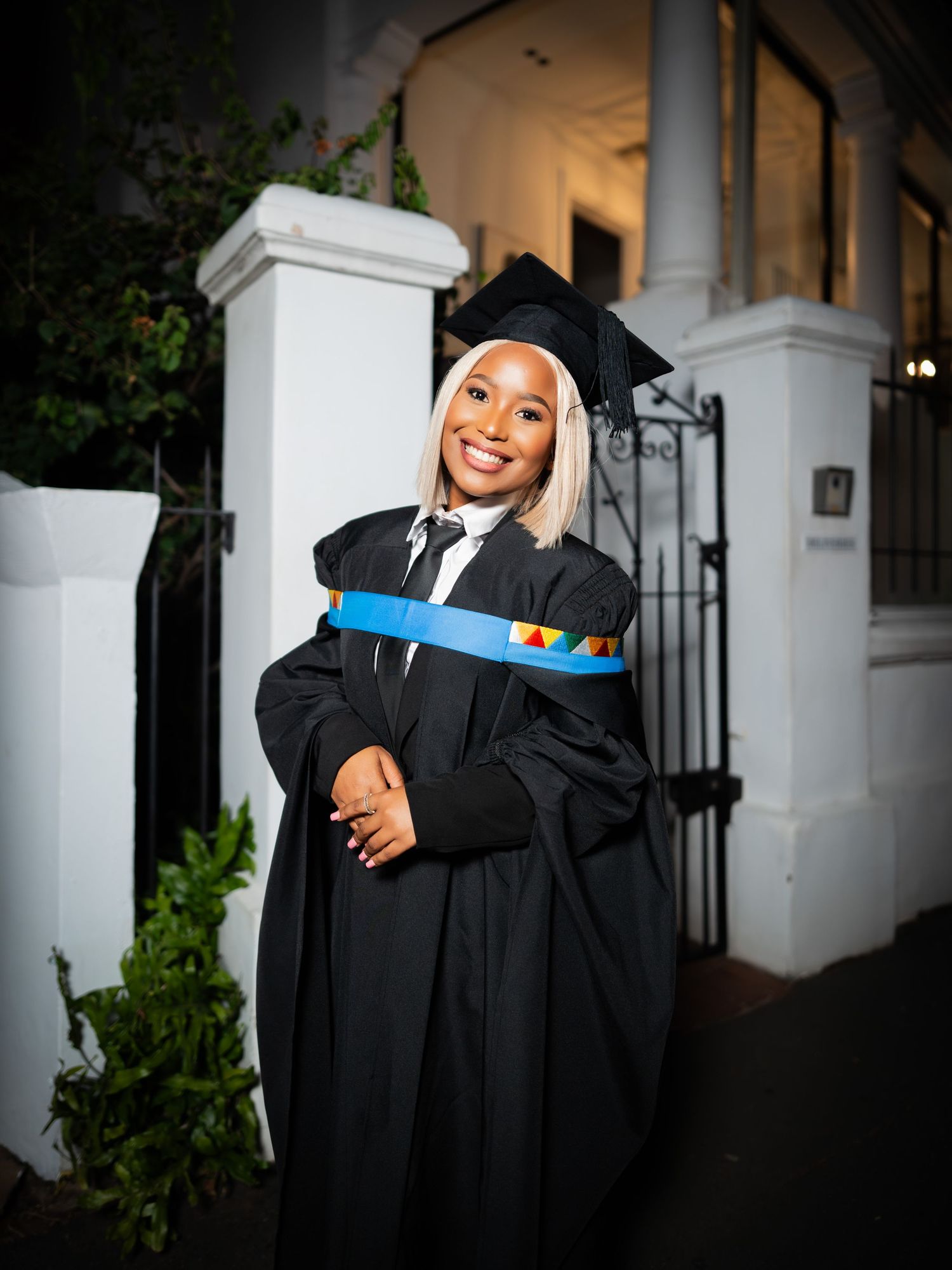 Smiling graduate in black cap and gown with colorful sash poses outdoors at night near white pillars.