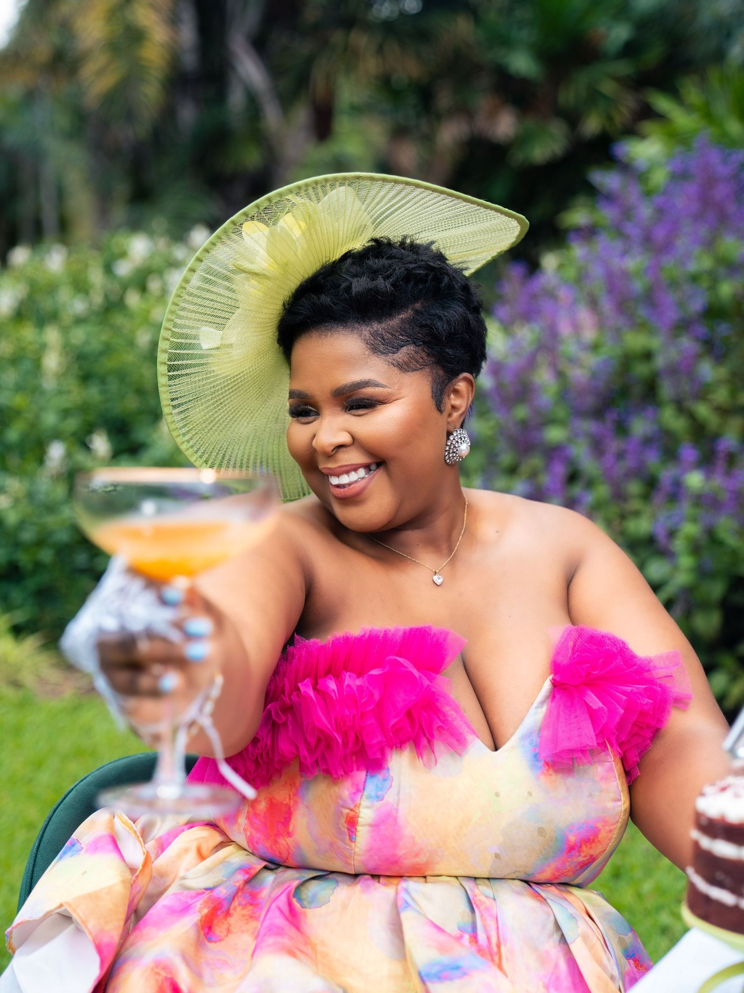 A joyful woman in a colorful strapless gown and green fascinator hat raises a cocktail glass outdoors.