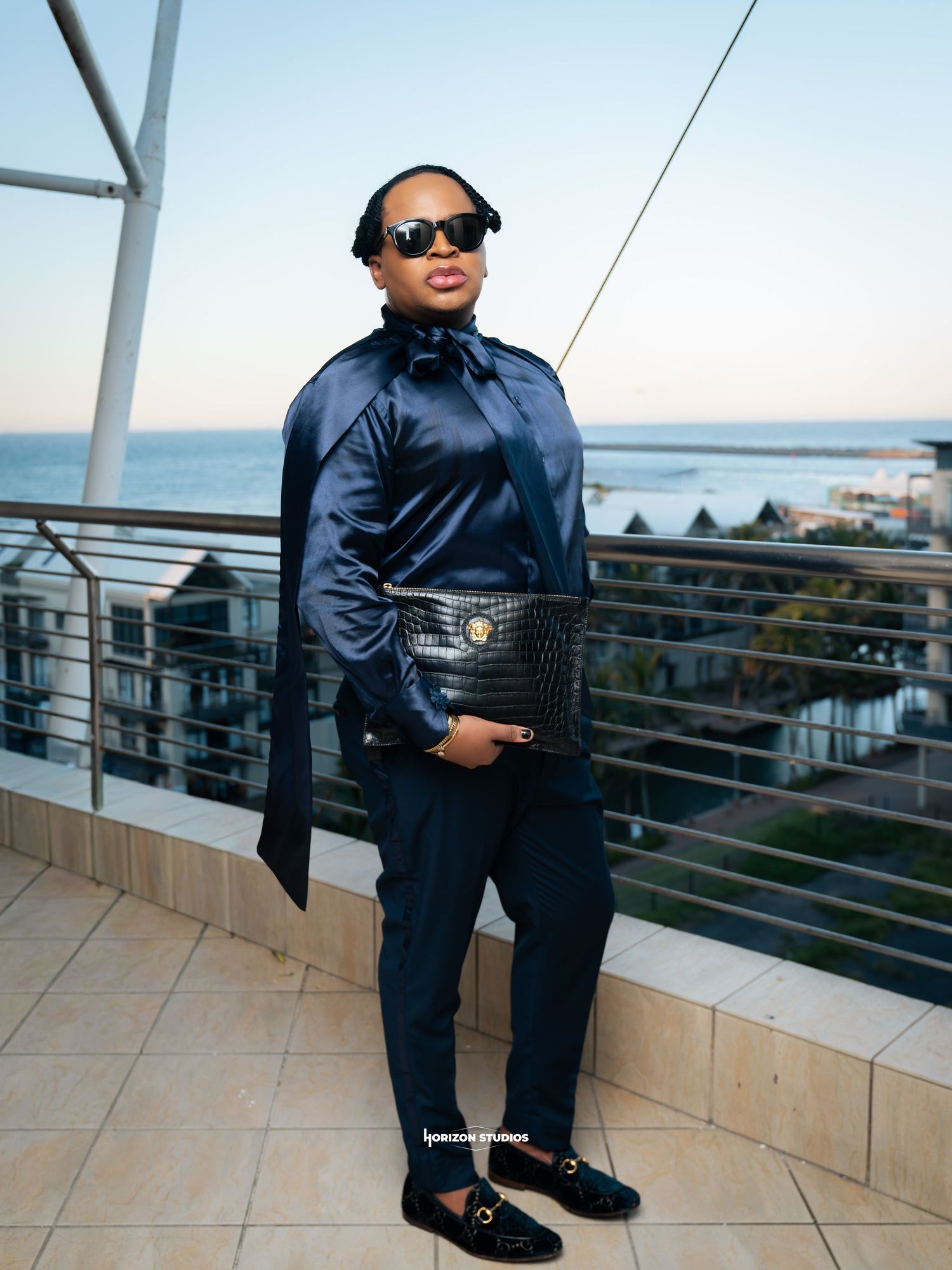 Stylish woman in navy satin blouse and trousers holding black croc clutch on seaside balcony.
