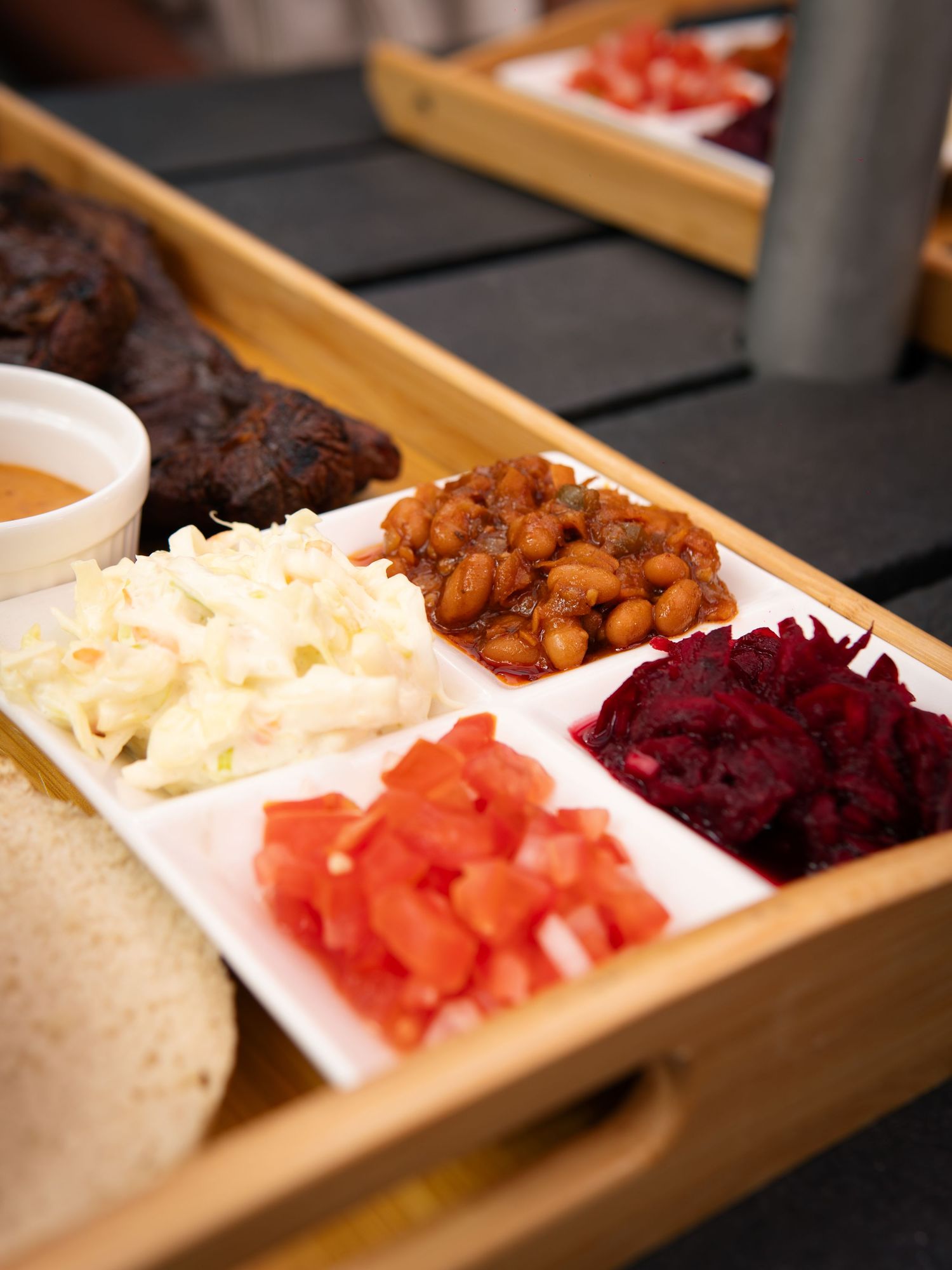 Wooden tray with white dish holding coleslaw, diced tomatoes, baked beans, and pickled beets beside BBQ meat and sauce.