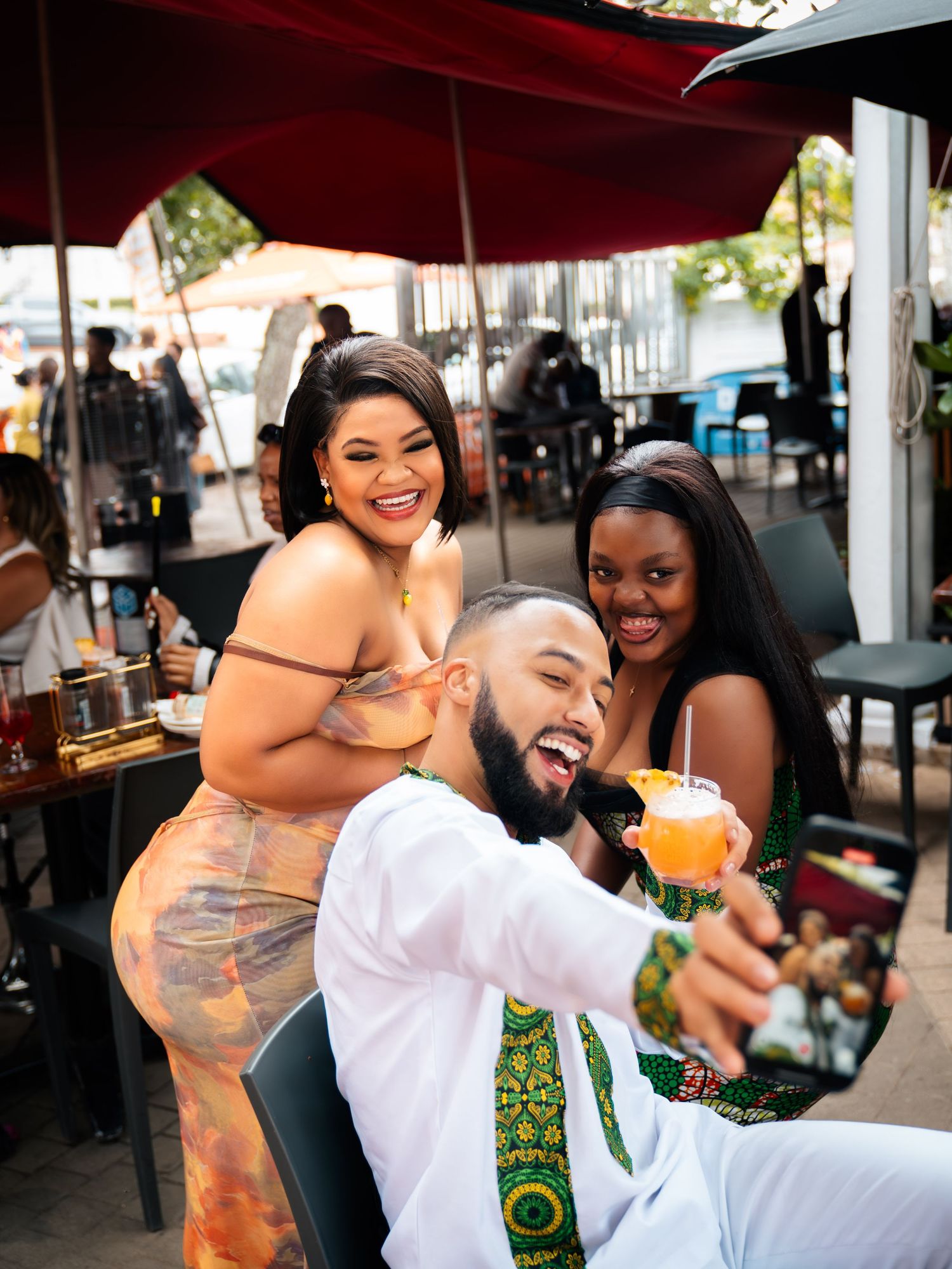 Three friends laughing and taking a selfie at an outdoor restaurant, holding tropical drinks in vibrant African print outfits.