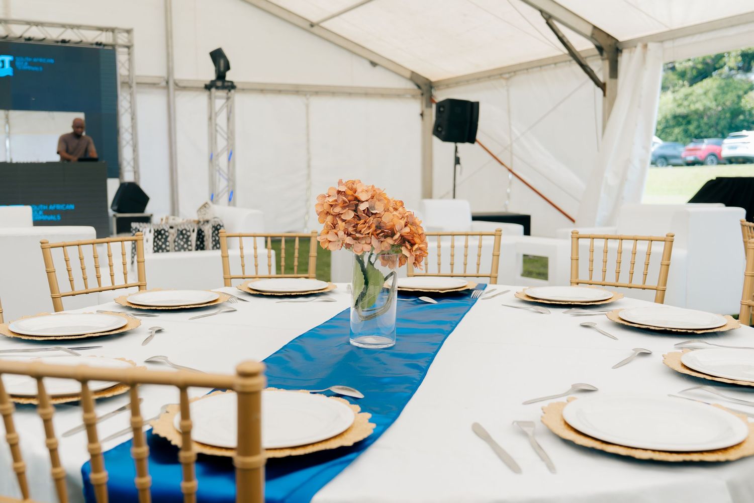 Elegant event tent table setting with gold Chiavari chairs, blue runner, peach floral centerpiece, and white plates on round table.