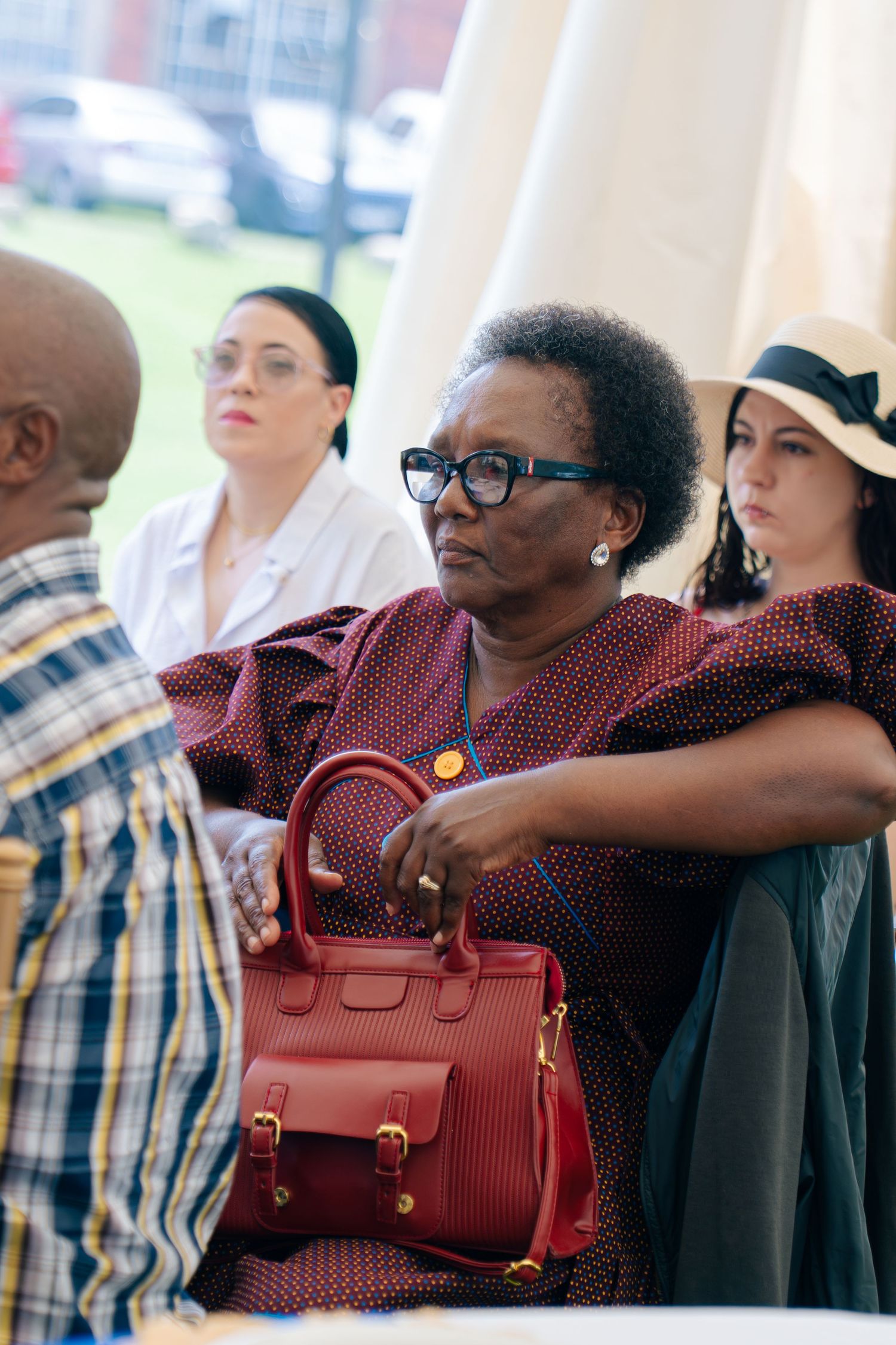 Elegant woman in burgundy dress with red handbag and glasses at outdoor gathering with diverse group.