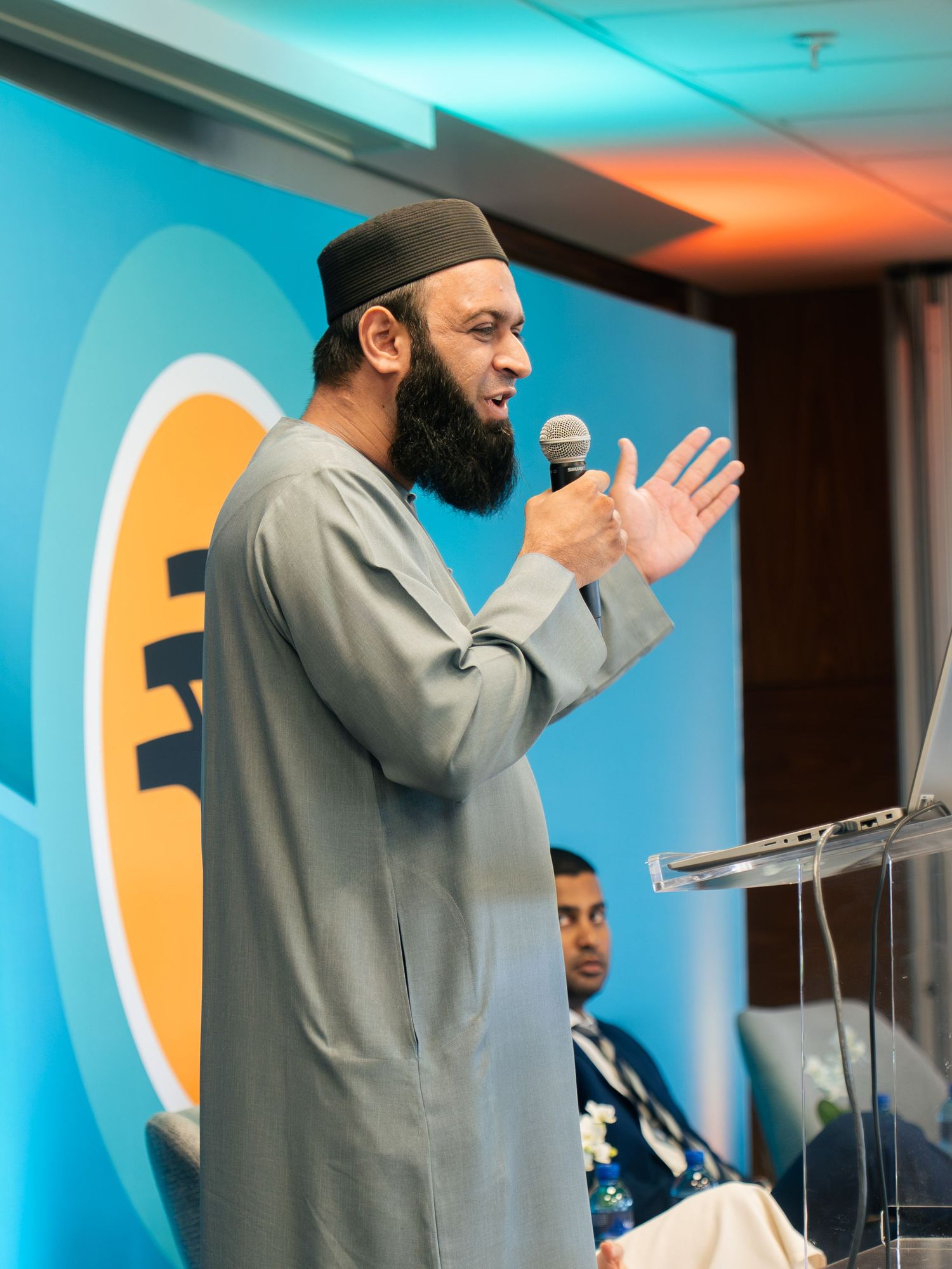Muslim speaker in gray thobe and kufi cap delivers speech at conference podium holding microphone.