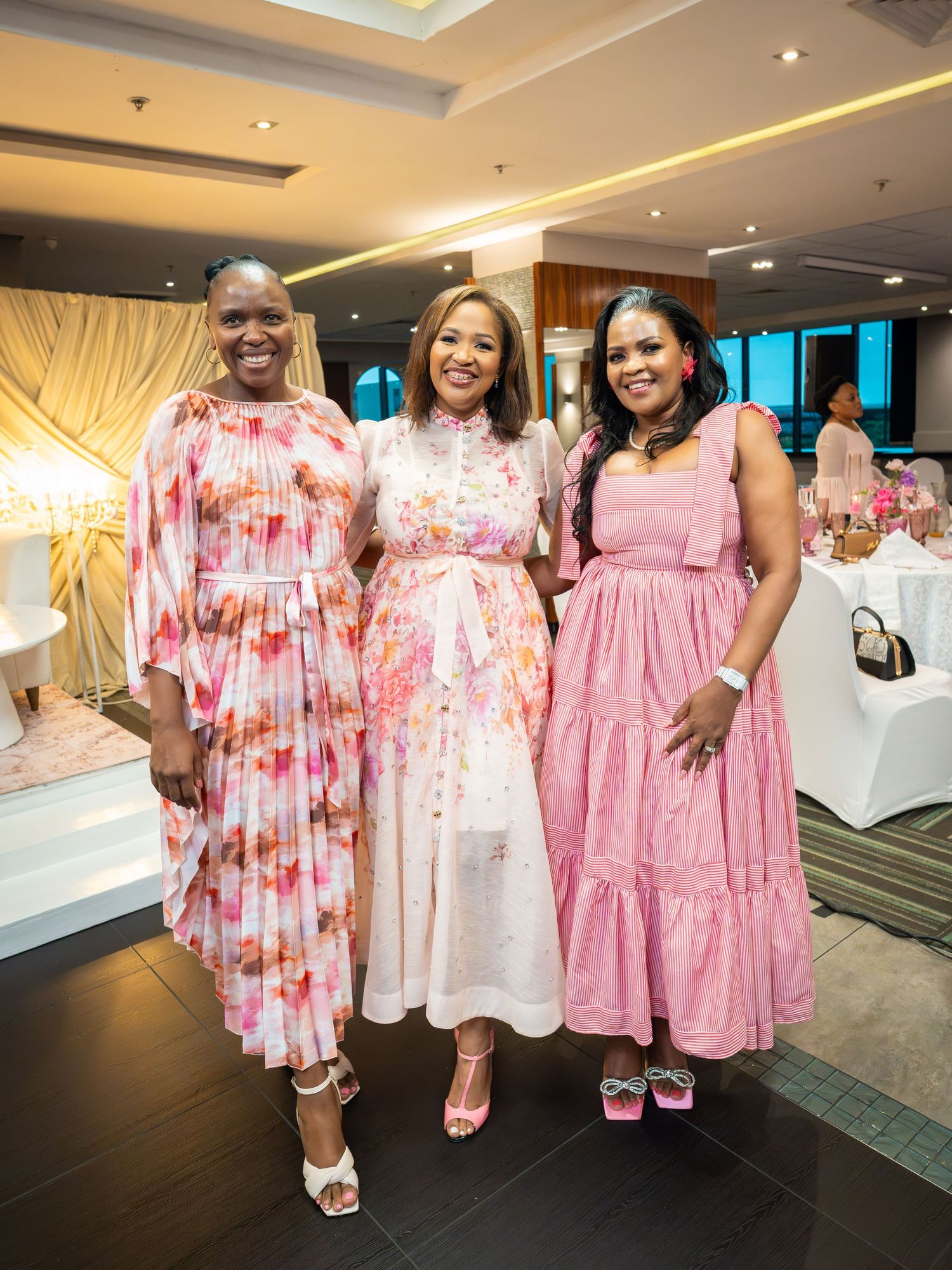 Three women in pink floral dresses pose together smiling at an elegant indoor event with white tablecloths.