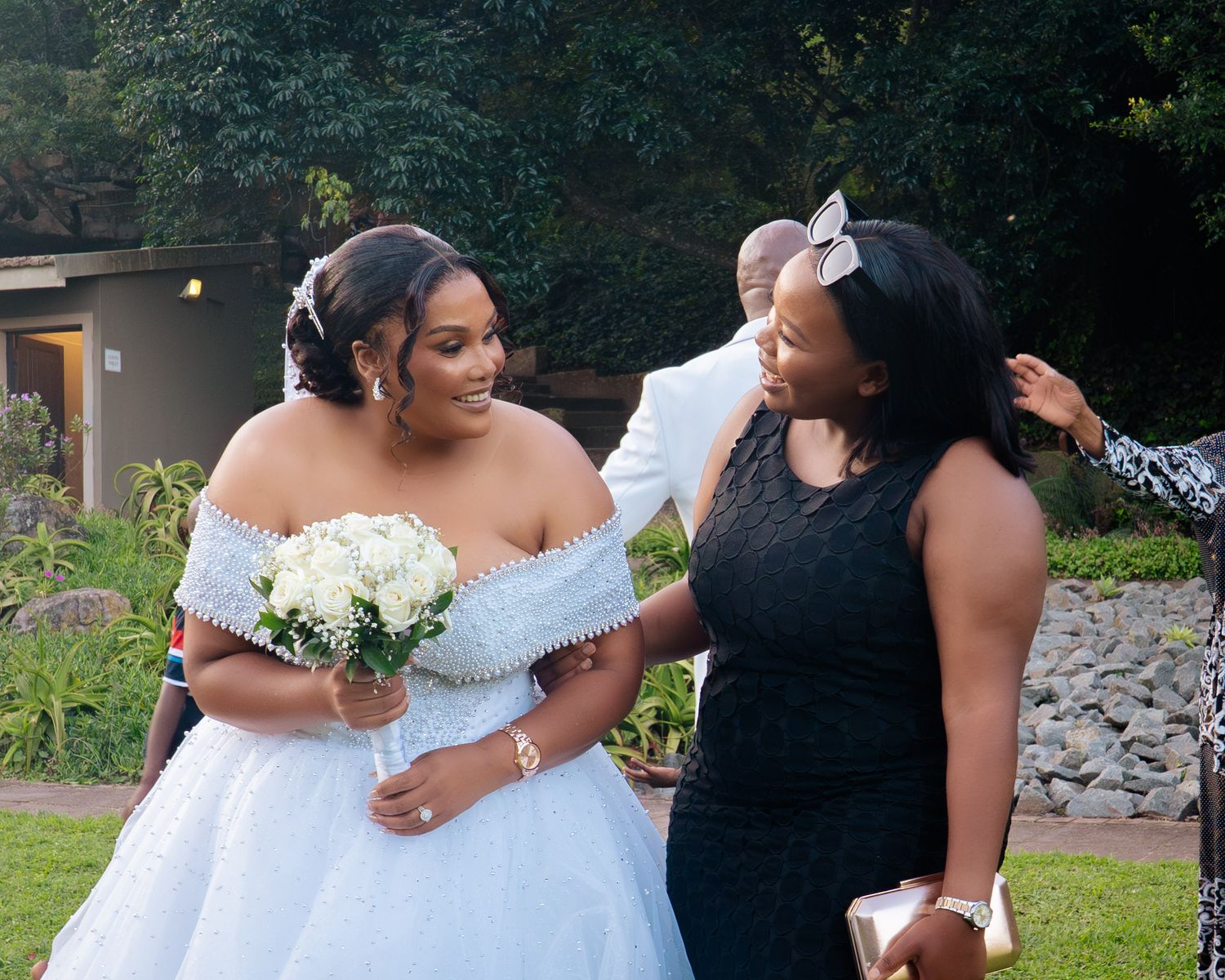 A bride in a white off-shoulder ball gown holds a bouquet while chatting with a guest in a black lace dress outdoors.