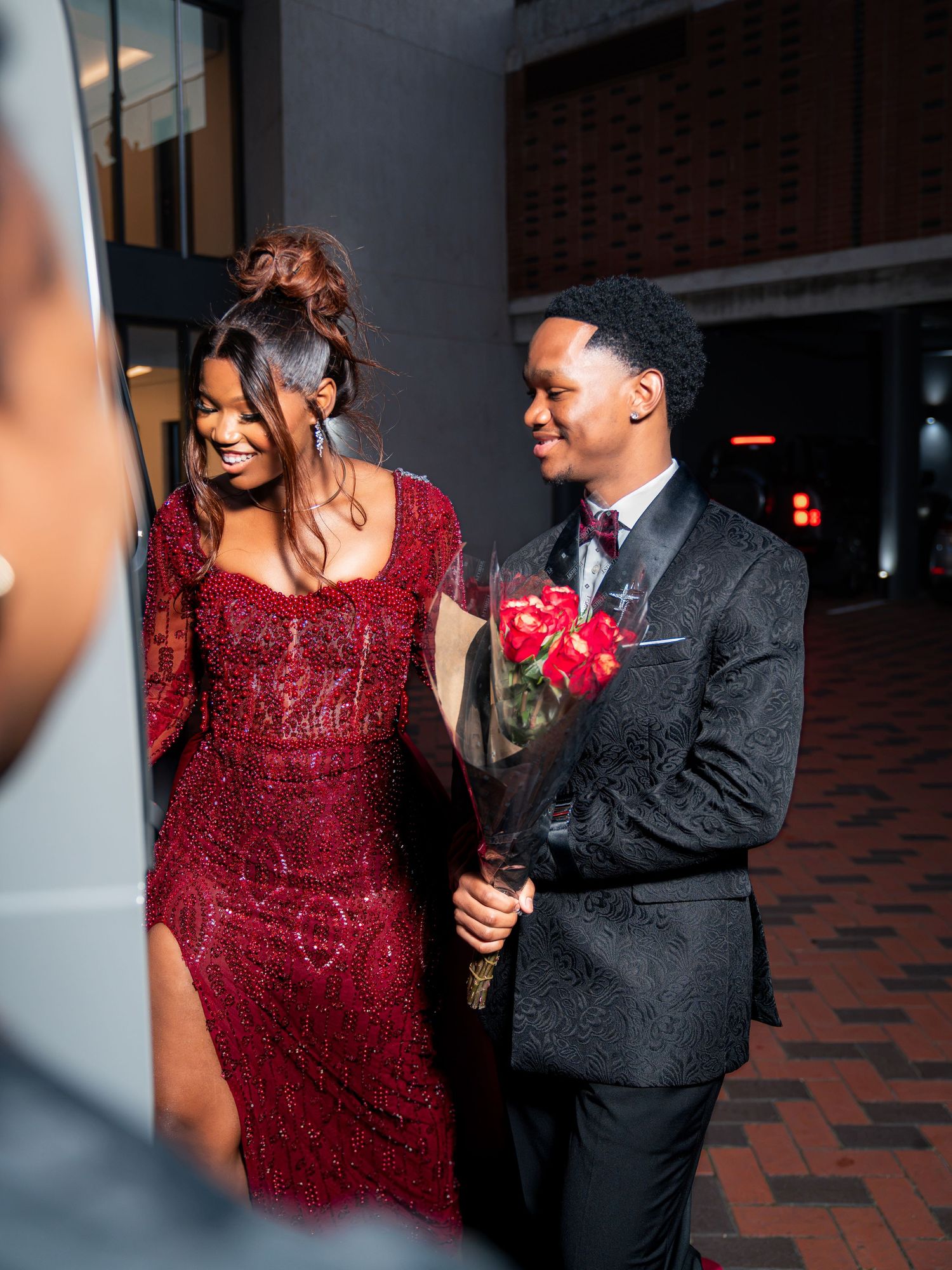 Couple in elegant red beaded gown and black tuxedo holding red roses at a formal night event.