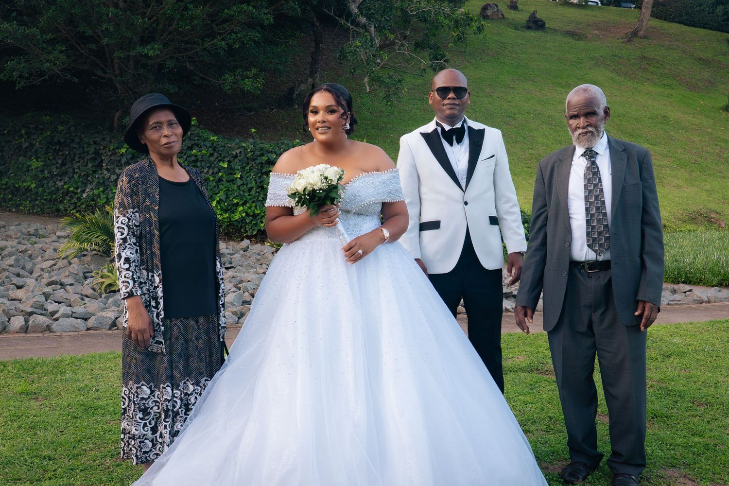 Bride in white ball gown poses with groom in tuxedo and two family members at an outdoor wedding ceremony.