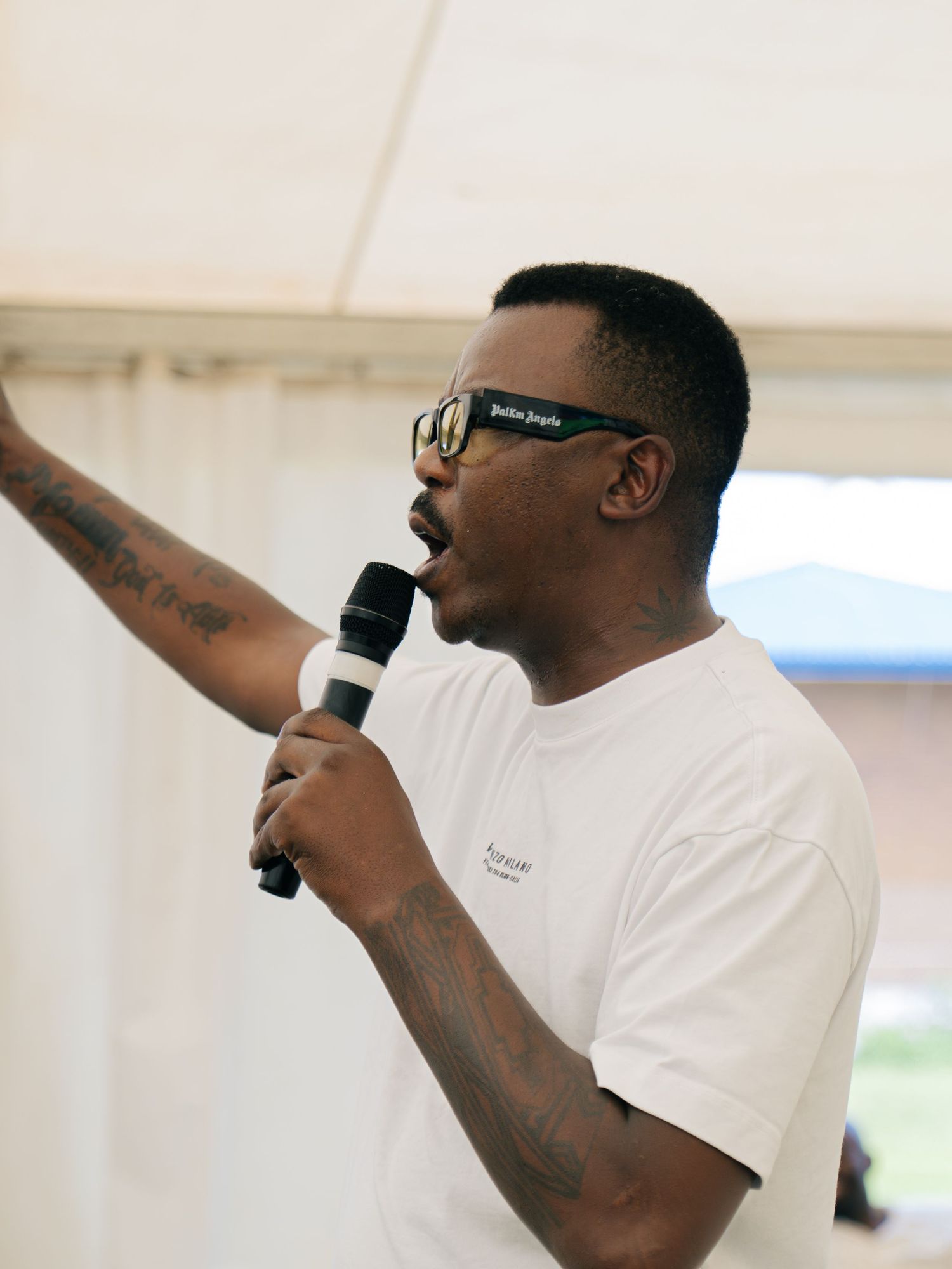 Man in white tee and sunglasses speaks into microphone with arm raised, showing tattoos at outdoor event.