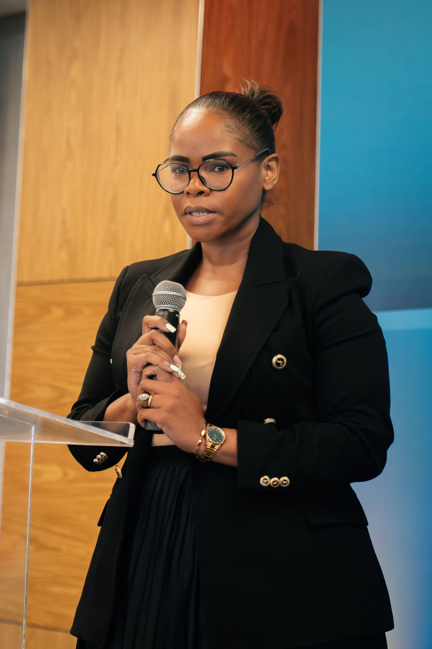 Woman in black blazer speaks at podium holding microphone, wearing glasses and gold accessories.