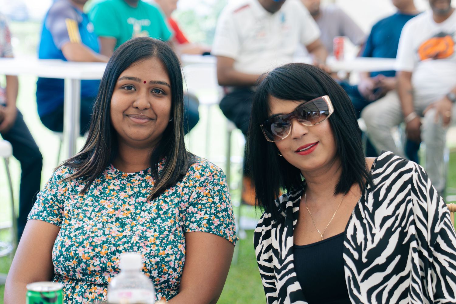Two women smiling at an outdoor event, one in a floral top and one in a zebra-print jacket with sunglasses.