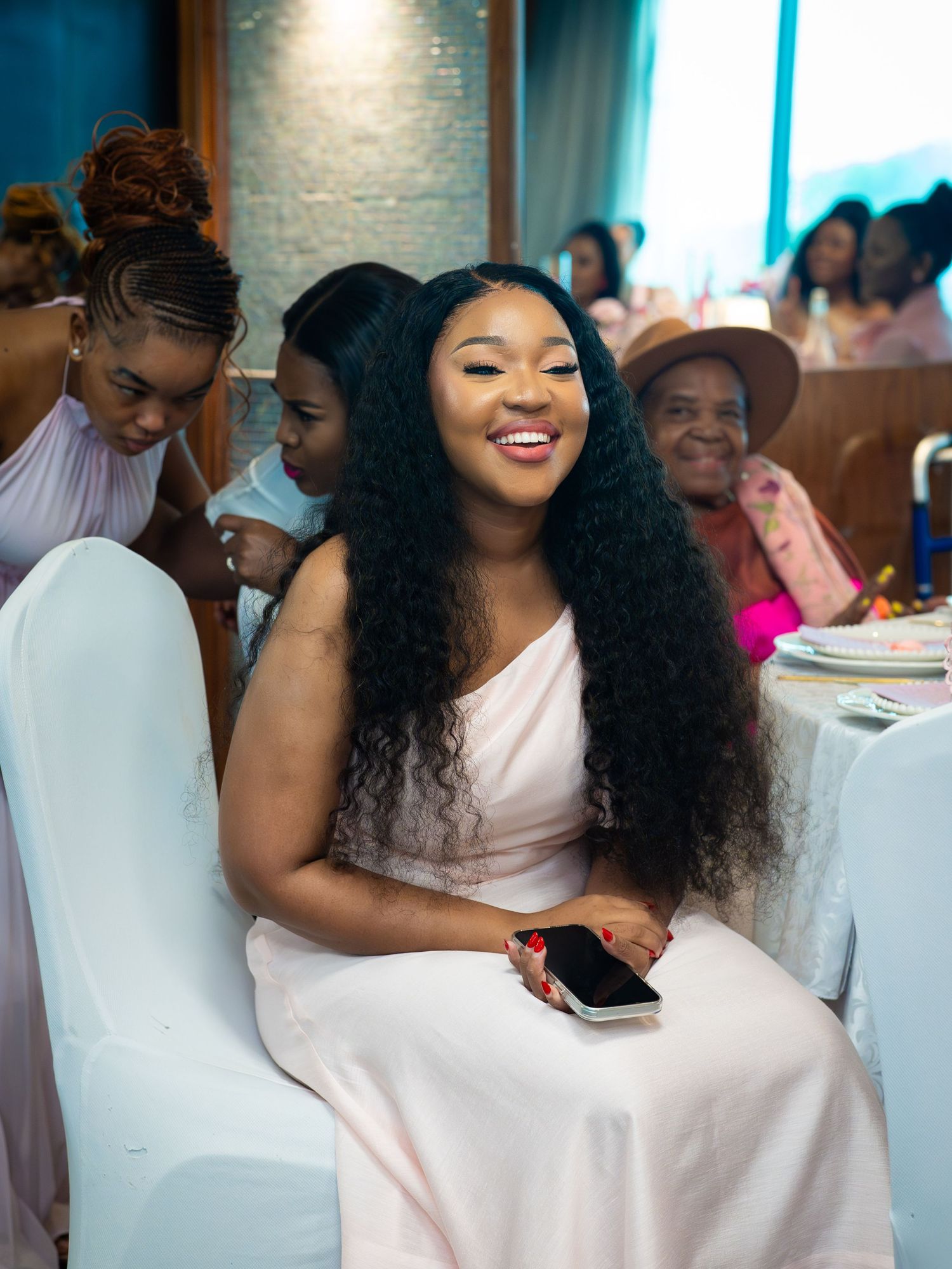 A smiling woman in a pink dress sits at an elegant event, holding a phone, with guests in the background.