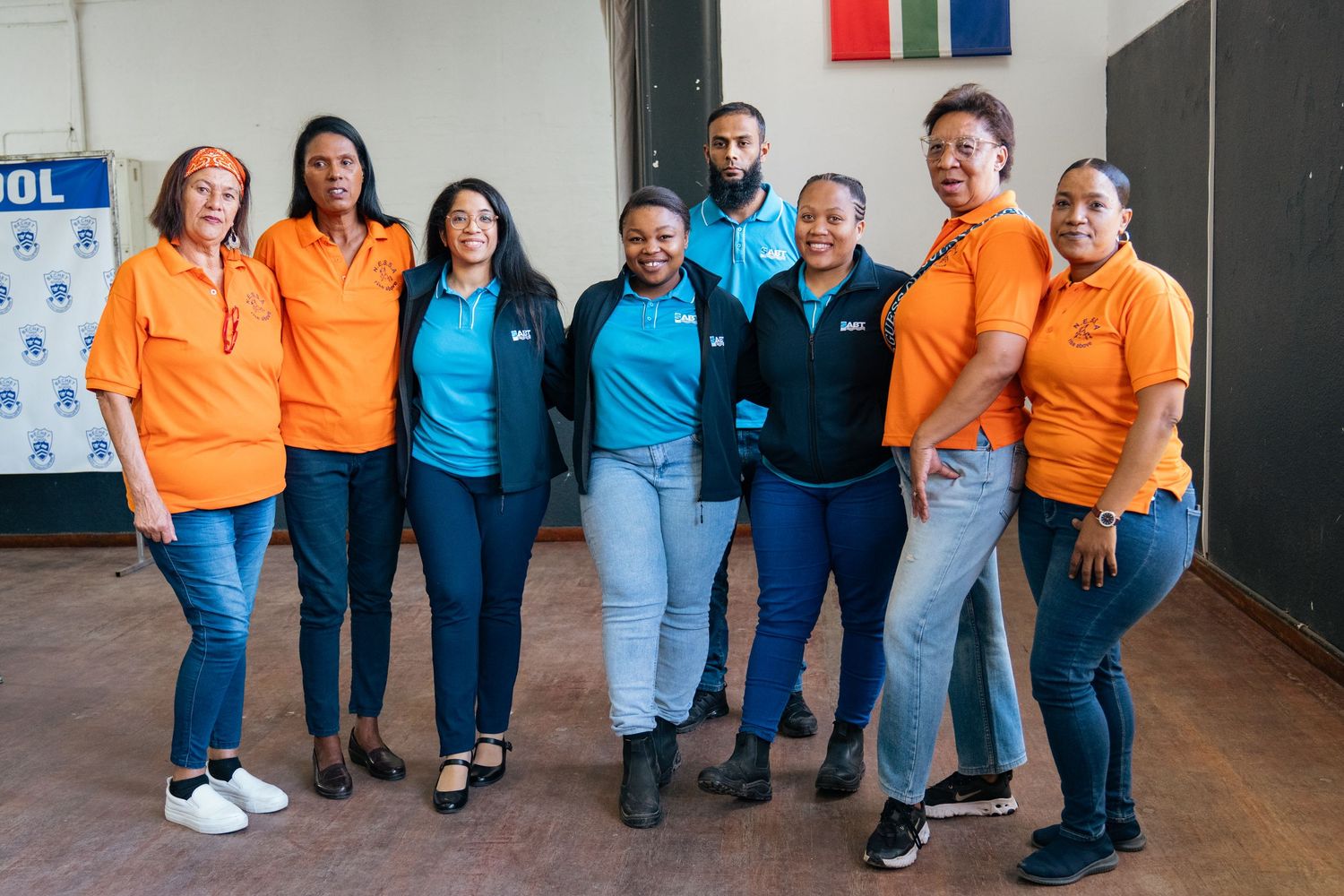 A diverse group of eight colleagues in orange and teal polo shirts posing together indoors for a team photo.