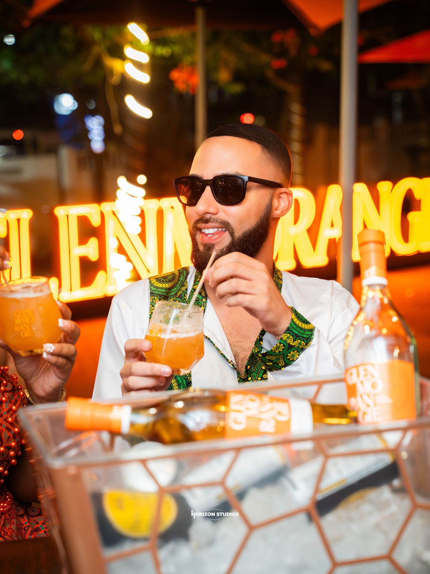 Man in sunglasses sipping orange cocktail at a vibrant nightclub with glowing neon signs.