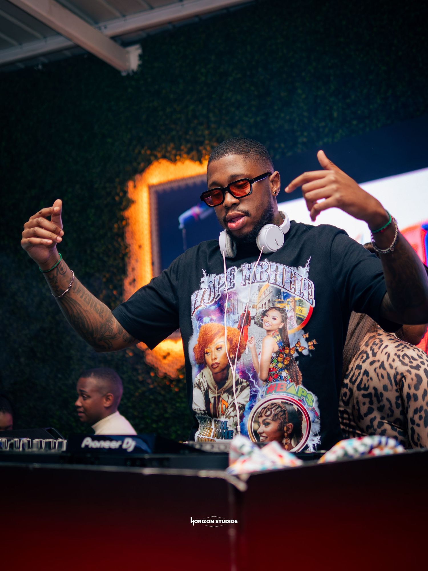 DJ performing at a nightclub event wearing headphones and a graphic tee, making hand gestures behind Pioneer DJ equipment.