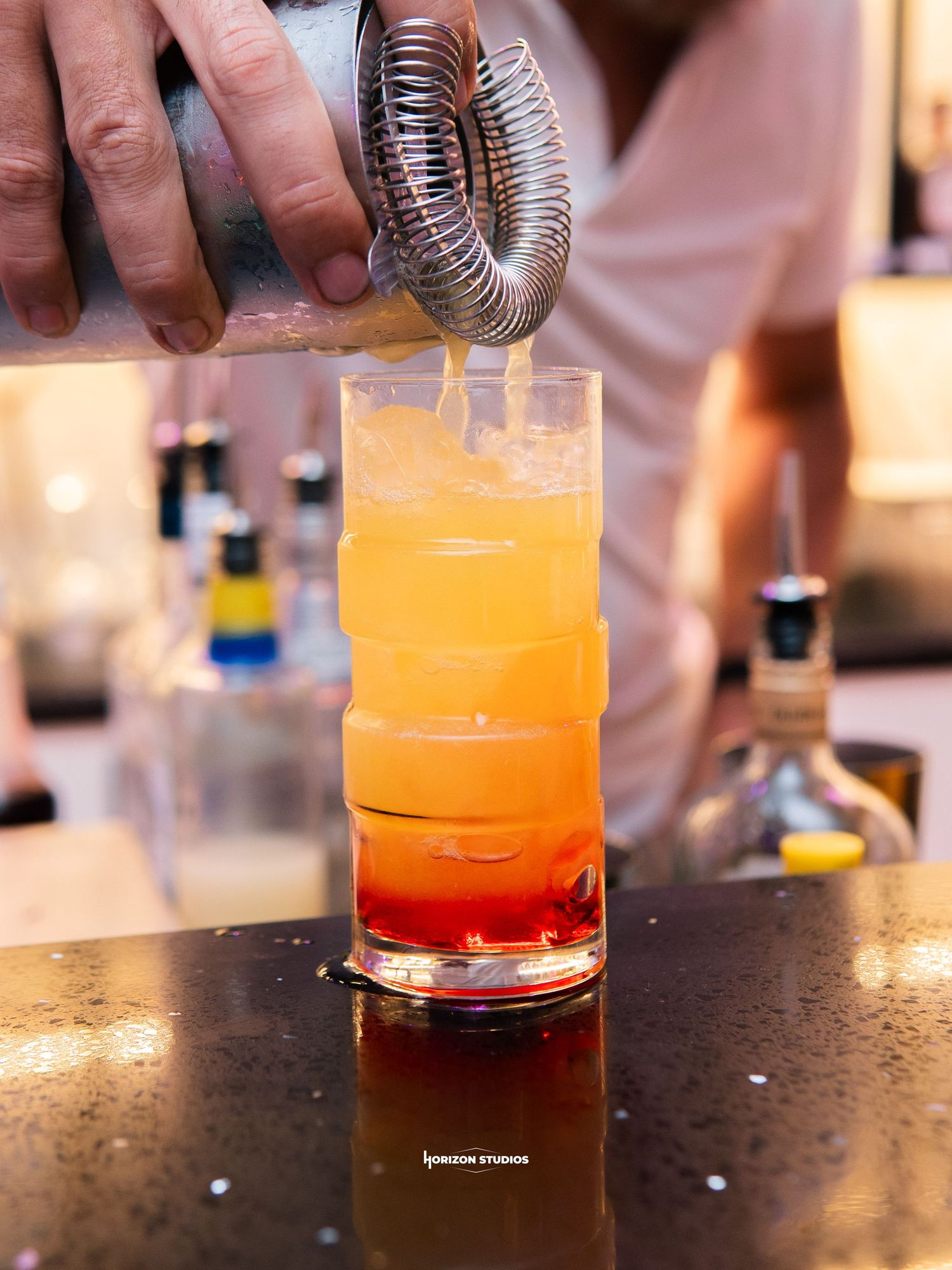 Bartender pouring a layered orange and red cocktail into a tall glass at a bar.