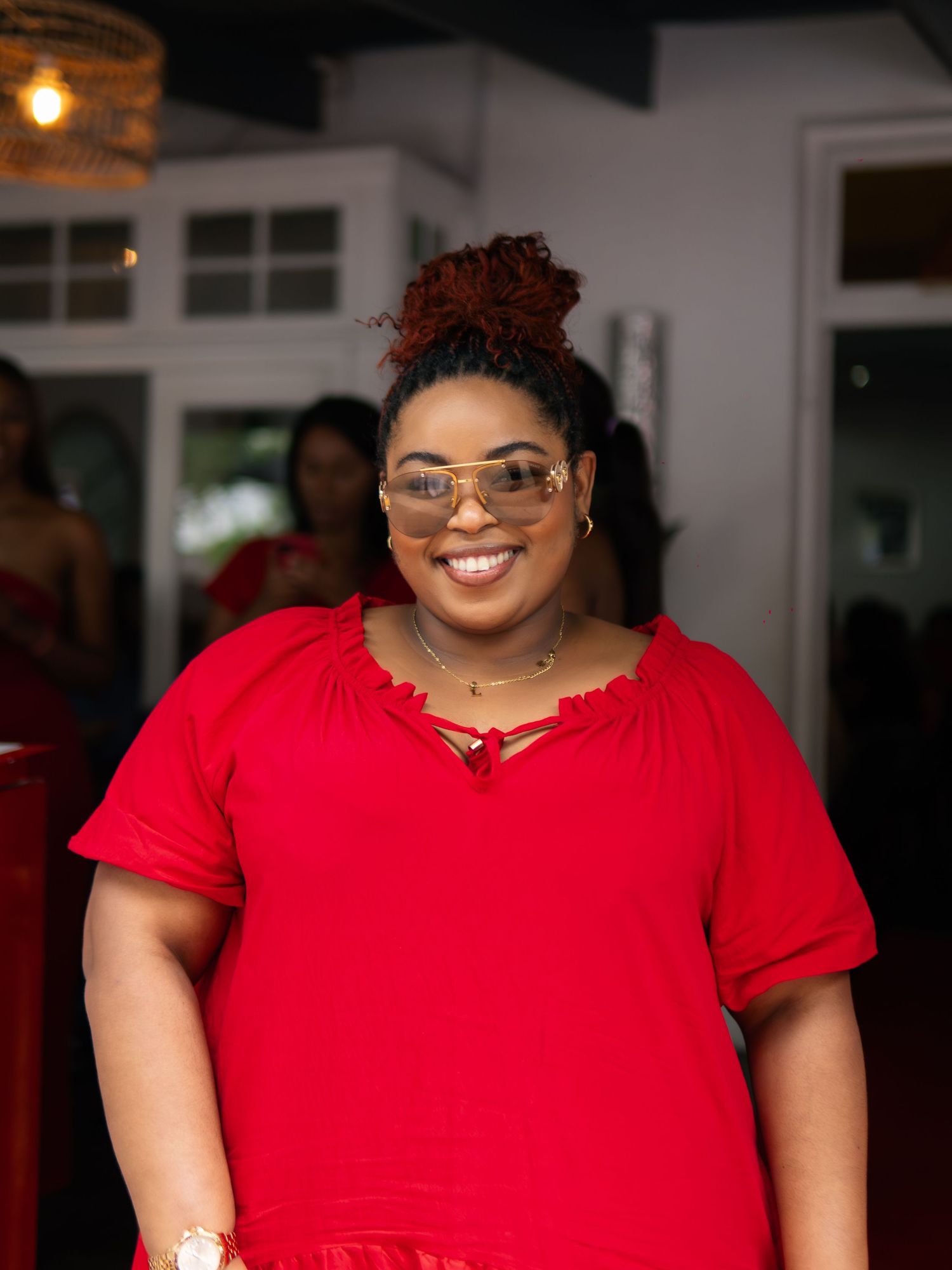 Smiling woman in red dress and aviator sunglasses with red updo hairstyle posing indoors at a social event.