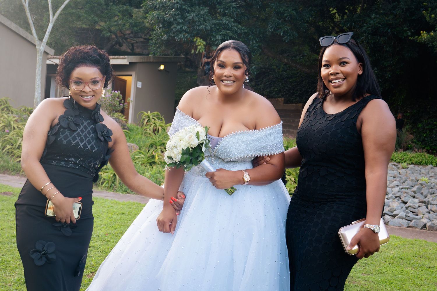 Bride in blue ball gown holds white bouquet flanked by two women in black dresses at outdoor wedding.