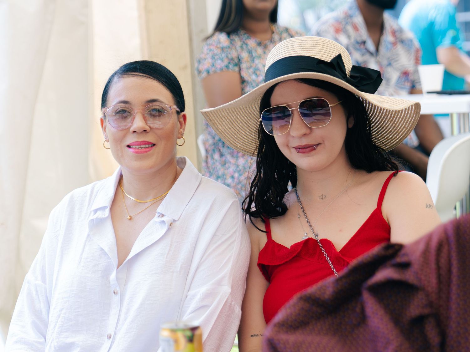 Two women smiling at an outdoor event, one in a white shirt with glasses, the other in a red dress and sun hat.