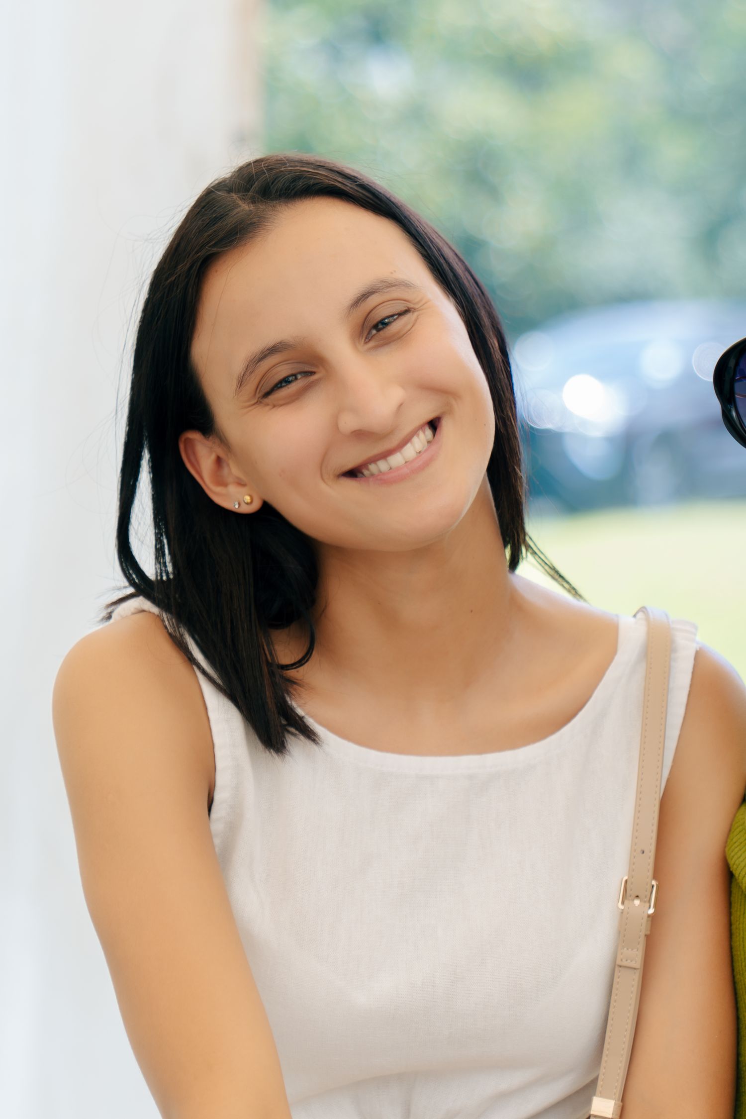 Smiling young woman with dark hair wearing a white sleeveless top, leaning against a wall outdoors.