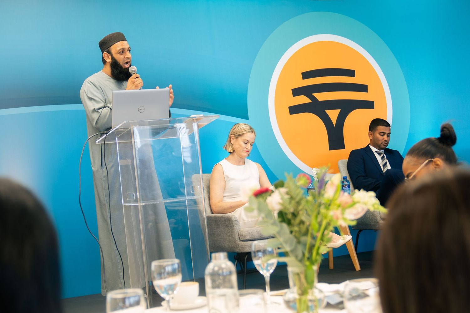 Man speaking at podium during formal event with seated panelists and orange tree logo backdrop.