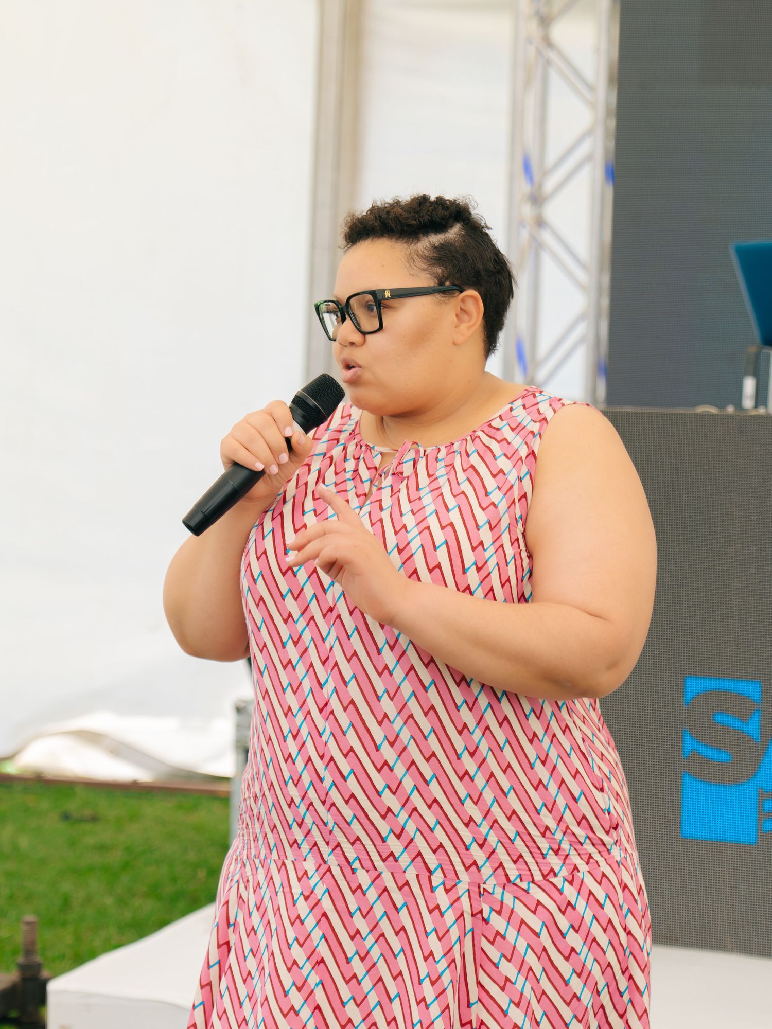 Woman in red patterned dress holding microphone speaks on outdoor stage wearing black glasses.