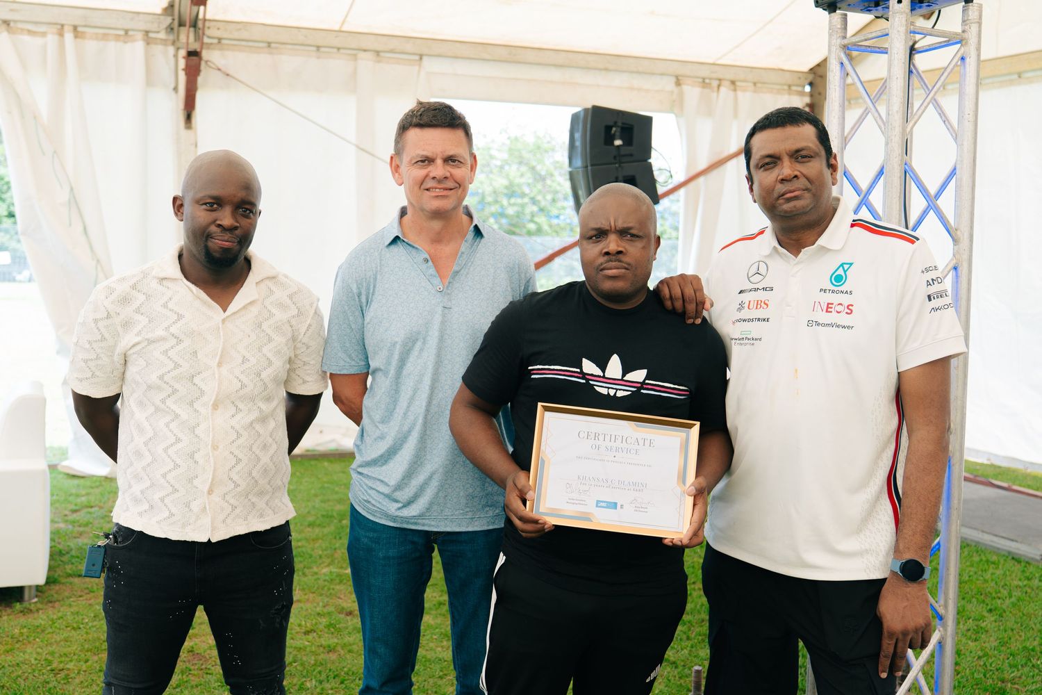 Four men pose together under a tent, one holding a framed certificate at an outdoor award ceremony.