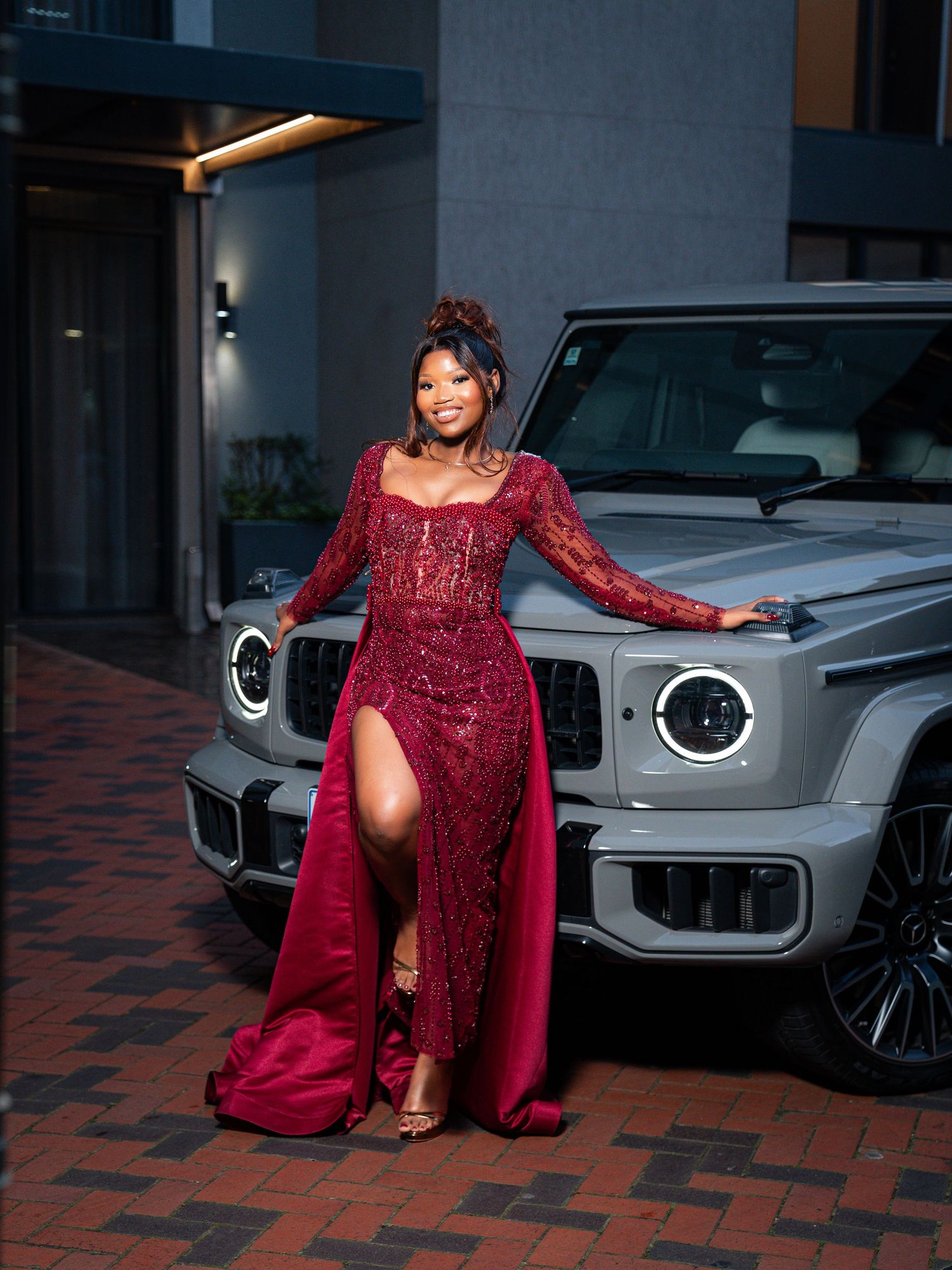 Woman in elegant burgundy lace gown with thigh slit posing beside Mercedes G-Wagon.