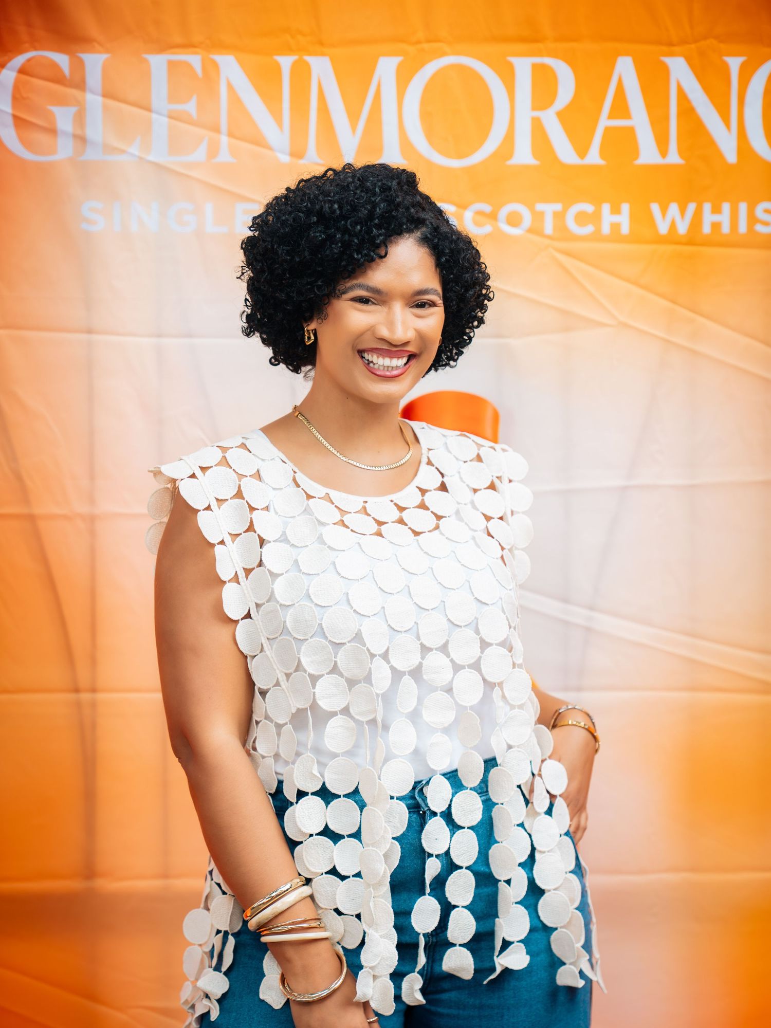 Woman smiling in white crochet top and teal pants at Glenmorangie Single Malt Scotch Whisky event.