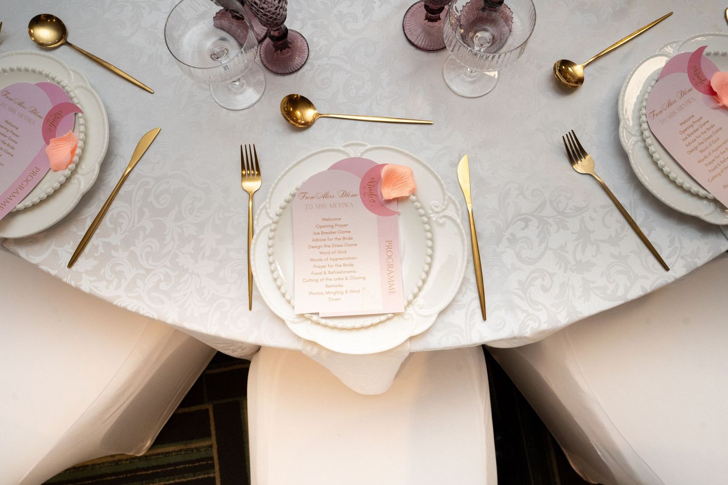 Elegant wedding table setting with gold cutlery, pearl-rimmed plates, pink menu cards, and purple glassware on white damask.