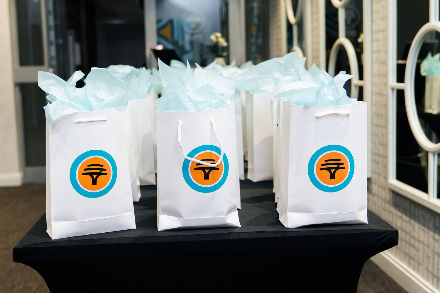 White gift bags with blue and orange logo filled with light blue tissue paper displayed on a black table.