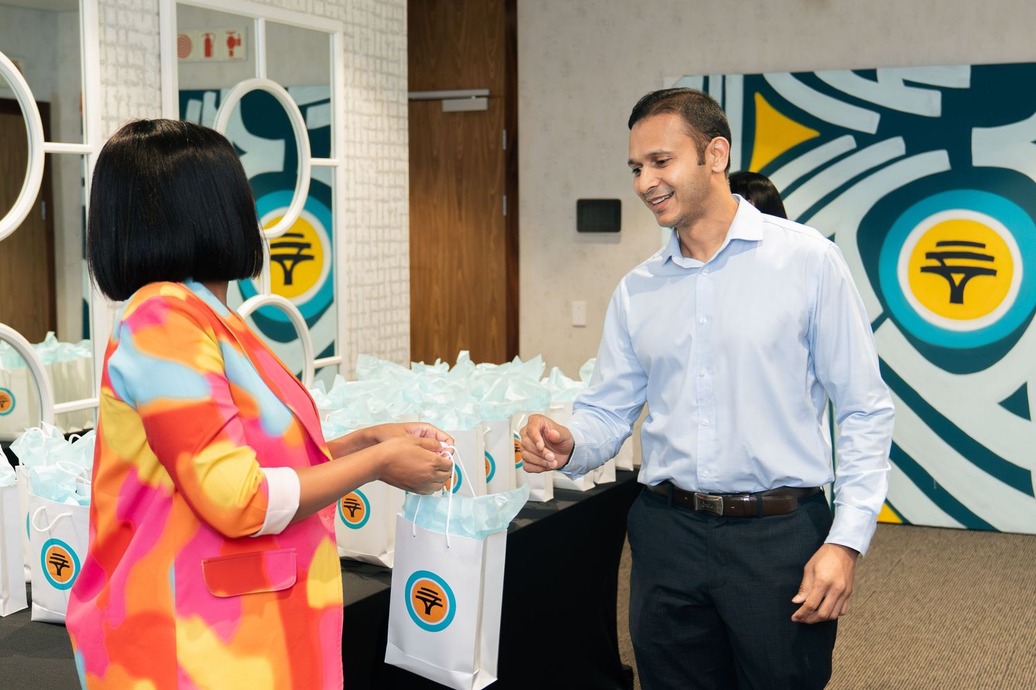 A man in a blue shirt exchanges gift bags with a woman in a colorful dress at a corporate event.